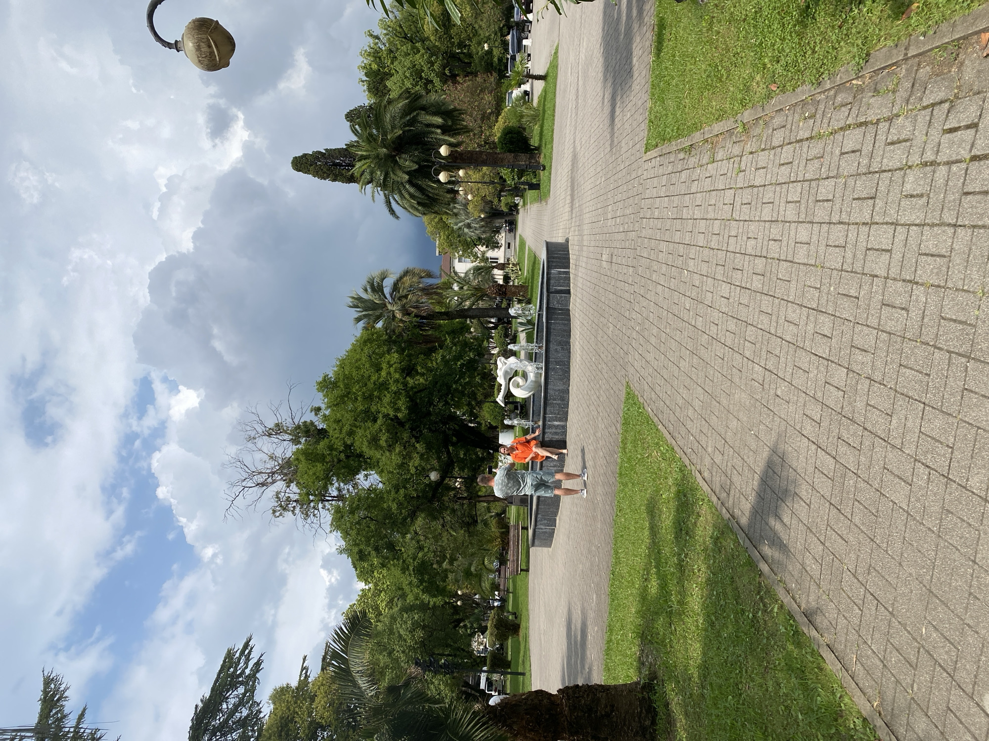 The image depicts a paved pathway in a park with lush green grass and trees, including palm trees. In the background, a fountain with white sculptures is active, with water spraying. Two people are visible near the fountain. A man, wearing a patterned t-shirt and shorts, stands facing away from the camera, holding a phone as if taking a picture or video. A woman, dressed in an orange dress, sits on the edge of the fountain. In the distance, there is a street with parked cars and buildings, and a lamppost with a distinctive lamp head is positioned in the upper right corner of the frame. The sky is partly cloudy with dramatic dark clouds present. The setting appears to be the central part of Sokhumi, Abkhazia, likely a public square or park area.