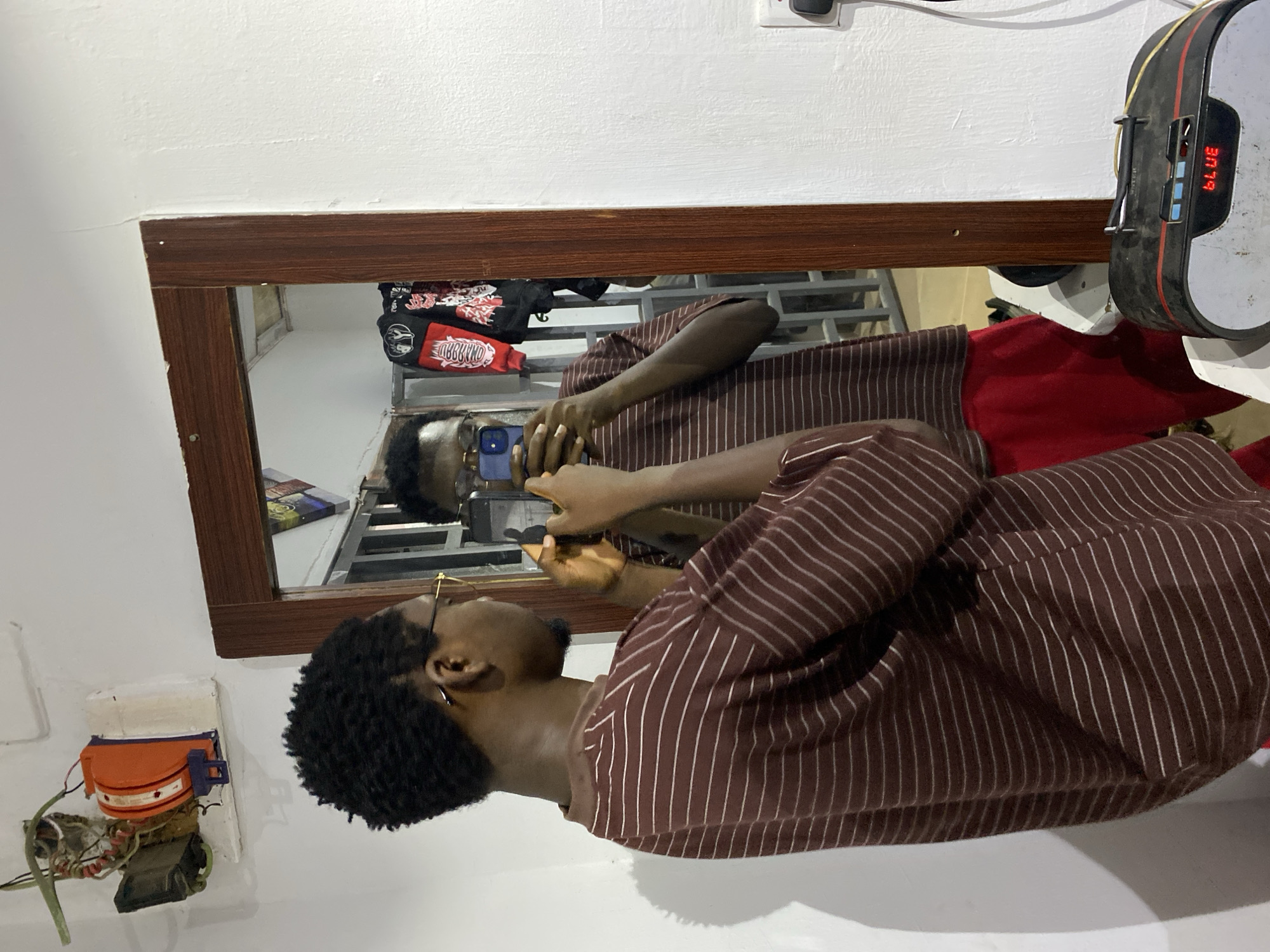 This image, captured in Jos, Nigeria, shows an indoor scene dominated by a large rectangular mirror with a dark wooden frame. The photo is rotated 90 degrees clockwise.
In the foreground, a young man with dark skin, short curly hair, and glasses is seen in profile, looking towards his reflection in the mirror. He is wearing a brown shirt with thin white vertical stripes. His reflection, seen from the front, occupies the center of the mirror. In the reflection, he is holding a blue-backed smartphone with both hands near his face, possibly taking a picture or adjusting his appearance. Behind his reflection, a metal shelving unit with bags and other items is visible, including one bag with the mirrored text "OBBЯAND" on a red patch.
The walls are white with a textured finish. To the lower left of the mirror, an exposed electrical installation is visible, featuring an orange and purple plastic unit with various wires (green, red, brown) and metal components. To the upper right of the mirror, a black and grey electronic device with red and blue accents is mounted on the wall. This device has a red LED display that clearly shows the word "BLUE" and a numerical reading, possibly "3079" or "30.79". The overall lighting suggests an indoor setting during the day or a well-lit room.
