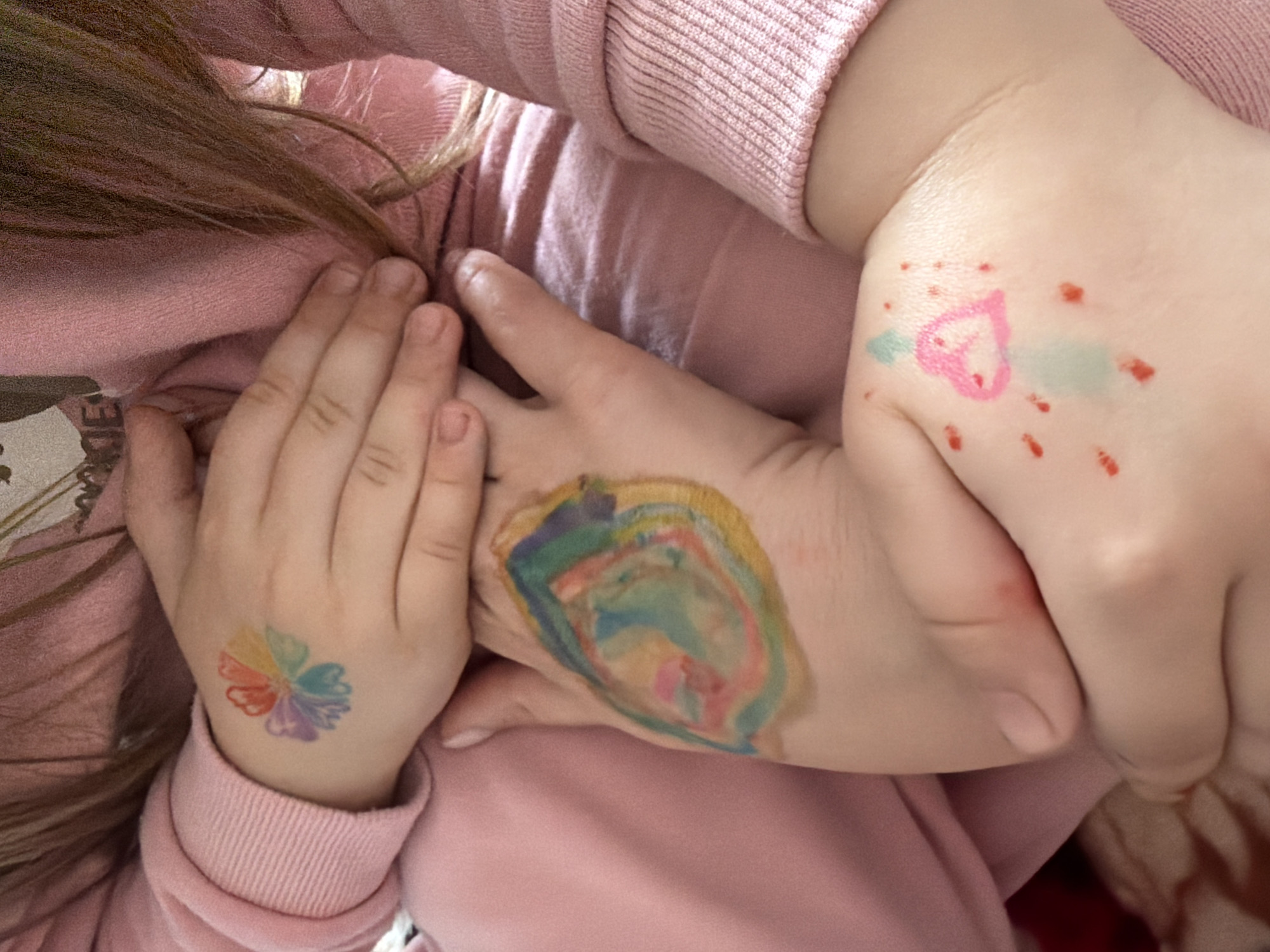 The image shows a close-up of a child's hands with colorful drawings and what appears to be a temporary tattoo on the back of the left hand. The child is wearing a pink long-sleeved shirt. The drawings on the right hand include a heart shape and scattered red dots, with a light blue smudge. The temporary tattoo on the left hand is a multi-colored flower. The background is out of focus and appears to be part of the child's clothing. The image does not provide any information or context that would allow for its location to be identified as Valozhyn, Belarus.