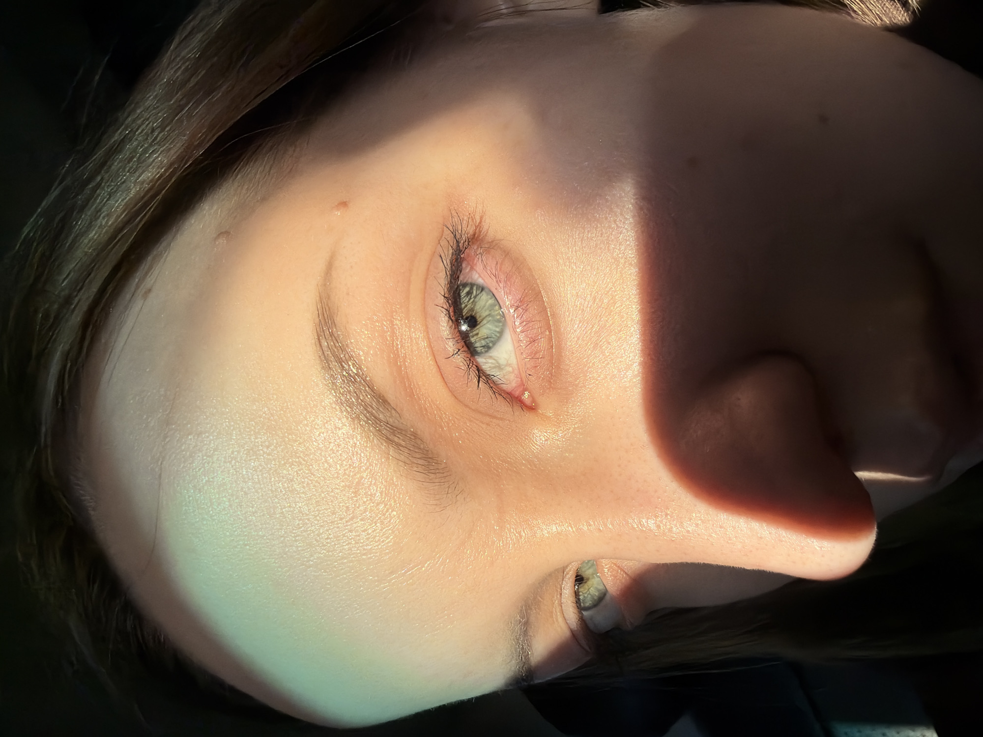 A close-up, angled shot of a person's face, primarily showcasing the eyes, nose, and forehead. The skin has a fair tone and exhibits visible pores. One eye is prominently featured, displaying a green iris, eyelashes, and surrounding skin. A prominent shadow falls across the face, creating a distinct contrast between light and dark areas. The subject has brown hair. The presence of the city of Minino, Russia, is not detectable within the image. The environment is otherwise not discernible.