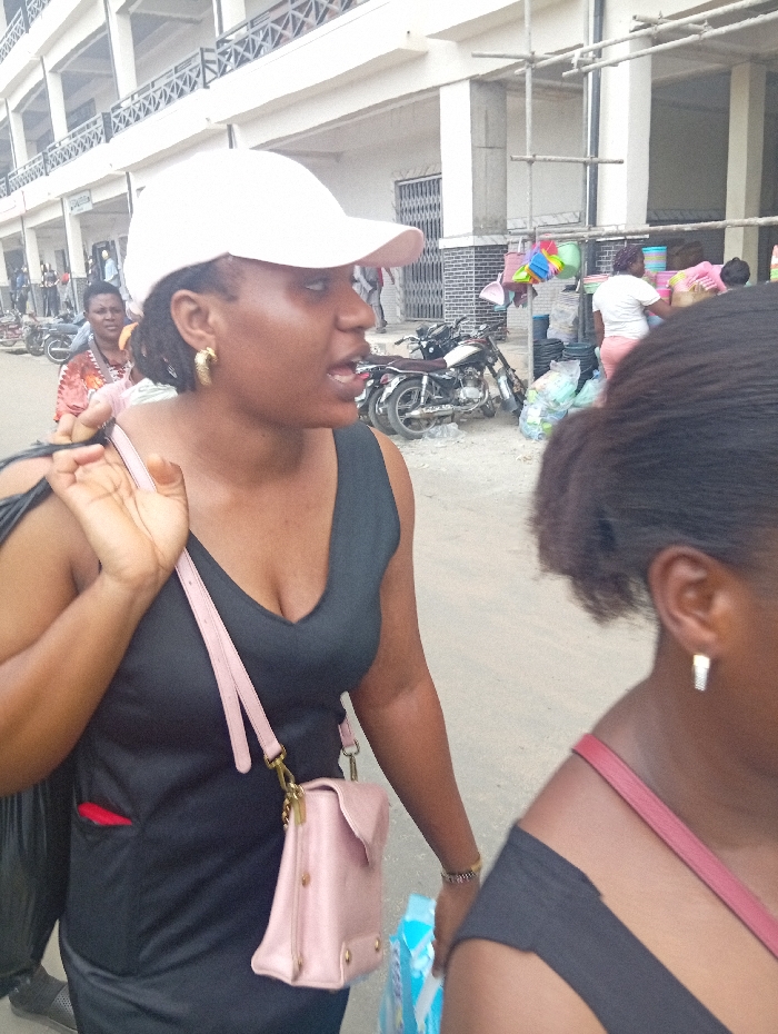 This image presents a daytime urban street and market scene in Ura Nta Umuarandu, Nigeria.
In the foreground, an adult Black female is depicted in profile, facing towards the right. She wears a light pink baseball cap, a black V-neck sleeveless top, and gold-colored hoop earrings. A light pink cross-body bag with gold-tone hardware is slung over her left shoulder, and a black bag strap is also visible on her left shoulder. Her mouth is slightly open. A red object is partially visible in a pocket on her left side. Her right hand carries a blue and white packaged item, and a bracelet is visible on her right wrist. To her immediate right, a partial view of another adult Black female shows a black sleeveless top and a reddish-pink shoulder strap, with a silver-colored stud earring visible on her ear.
The midground features a busy street with several motorbikes parked on a mixed paved and unpaved surface. To the right, an open-air market stall displays numerous brightly colored plastic household items, including buckets and basins, suspended from a metal frame. Multiple individuals, including adult females in light-colored tops, are visible interacting with this market display. Bags of various goods are present on the ground near the stall.
In the background, multi-story buildings with balconies and barred windows line the street, indicating commercial or residential structures. Scaffolding is visible against one of the buildings on the right.