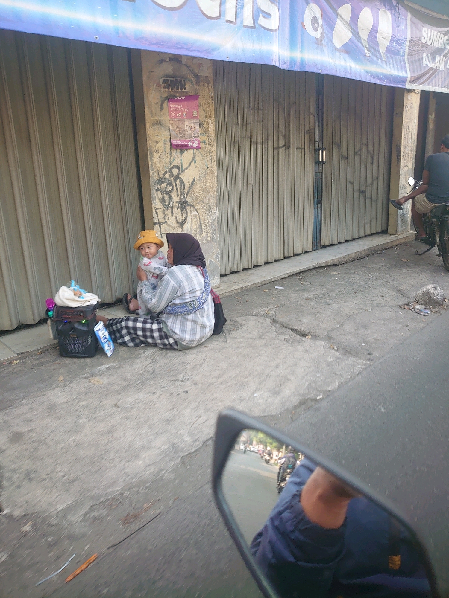 A woman wearing a hijab and a plaid shirt sits on a concrete sidewalk next to a corrugated metal storefront. She holds a baby wearing a yellow hat. A black basket containing a white cloth and a pink object is beside her. A man on a motorcycle is visible to the right, near the storefront entrance. The scene is likely on a street in Depok, Indonesia, given the architecture and the presence of a motorcycle.