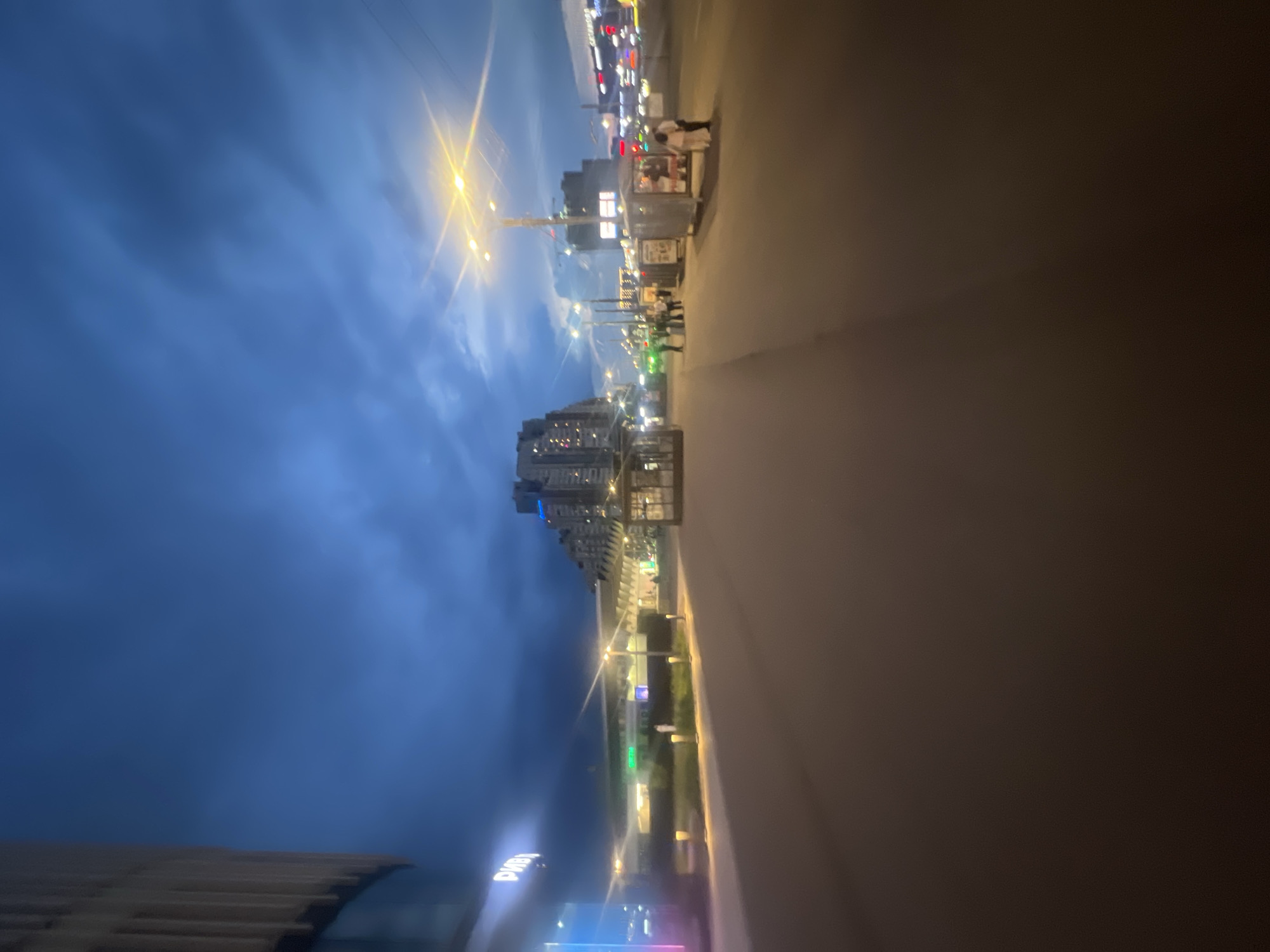 The image depicts an urban street scene at twilight or night in Saint Petersburg, Russia, specifically in the vicinity of the Riviera shopping mall, indicated by the partial Cyrillic text "РИВ" visible on the prominent modern building to the left. This building features a curved facade constructed with vertical light-colored panels and a glass lower section. The sky overhead is dark blue with discernible cloud formations. A wide, dark paved area, likely a pedestrian walkway or urban plaza, occupies the foreground and extends into the mid-ground.
The scene is extensively illuminated by artificial light sources. Multiple streetlights with bright yellow-orange output are positioned on tall poles throughout the mid-ground and background, casting radial light flares. In the mid-ground to the left, a series of lower structures with illuminated interiors are visible, surrounded by landscaped elements and small, upward-shining bollard lights. On the right side of the mid-ground, several kiosks or small commercial stalls are present. Two individuals, dressed in light-colored clothing, are standing near one of these kiosks. Additional indistinct human figures are scattered further along the street and in the background, appearing to be walking or standing. Multi-story buildings with illuminated windows form the distant backdrop, contributing to the overall urban lightscape.