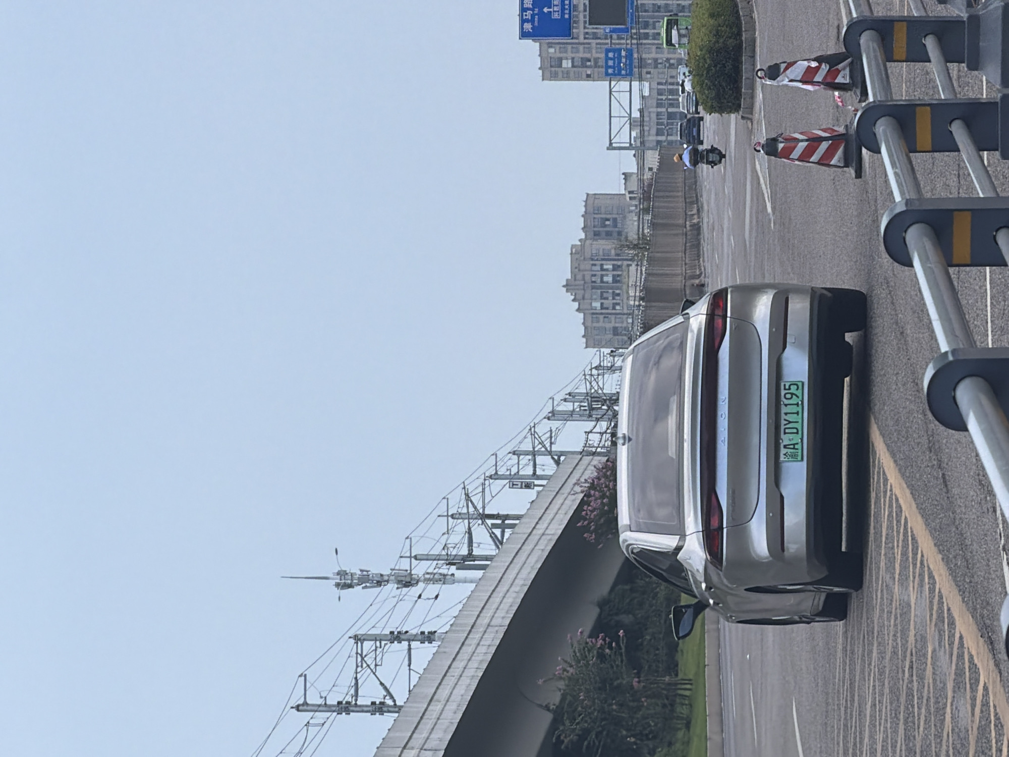 A silver sedan, license plate A·DY1195, is driving on an elevated highway in Jiangjin, China. The car is in the right lane, passing under a viaduct for an elevated railway system, which is visible overhead with its catenary wires and support structures. The background features multi-story buildings and a hazy sky. On the right side of the road, traffic cones are placed near a guardrail, and other vehicles, including a scooter and a bus, are visible further down the road. The environment appears to be an urban setting during the daytime.