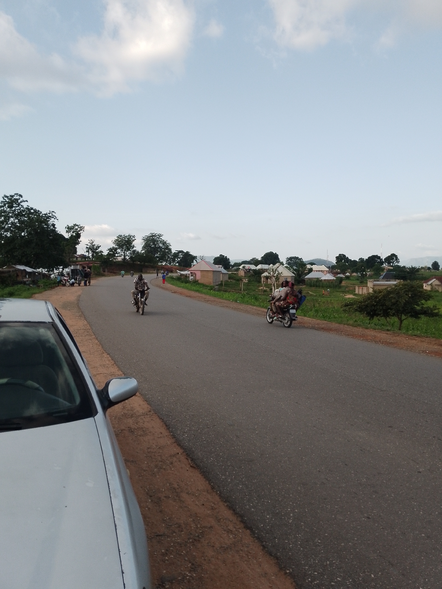 In the city of Serti, Nigeria, an overcast sky looms over a rural road. The road itself is paved with asphalt and curves to the right, bordered by a dirt shoulder. On the left, a silver car is parked, its side and side mirror visible. A group of people and motorcycles are gathered on the left side of the road. Further down the road, a man rides a motorcycle, and another motorcycle carries two passengers. The environment is characterized by scattered buildings, lush green vegetation, and trees. The scene depicts a typical day in a Nigerian village, with everyday activities taking place on the road and in the surrounding areas.