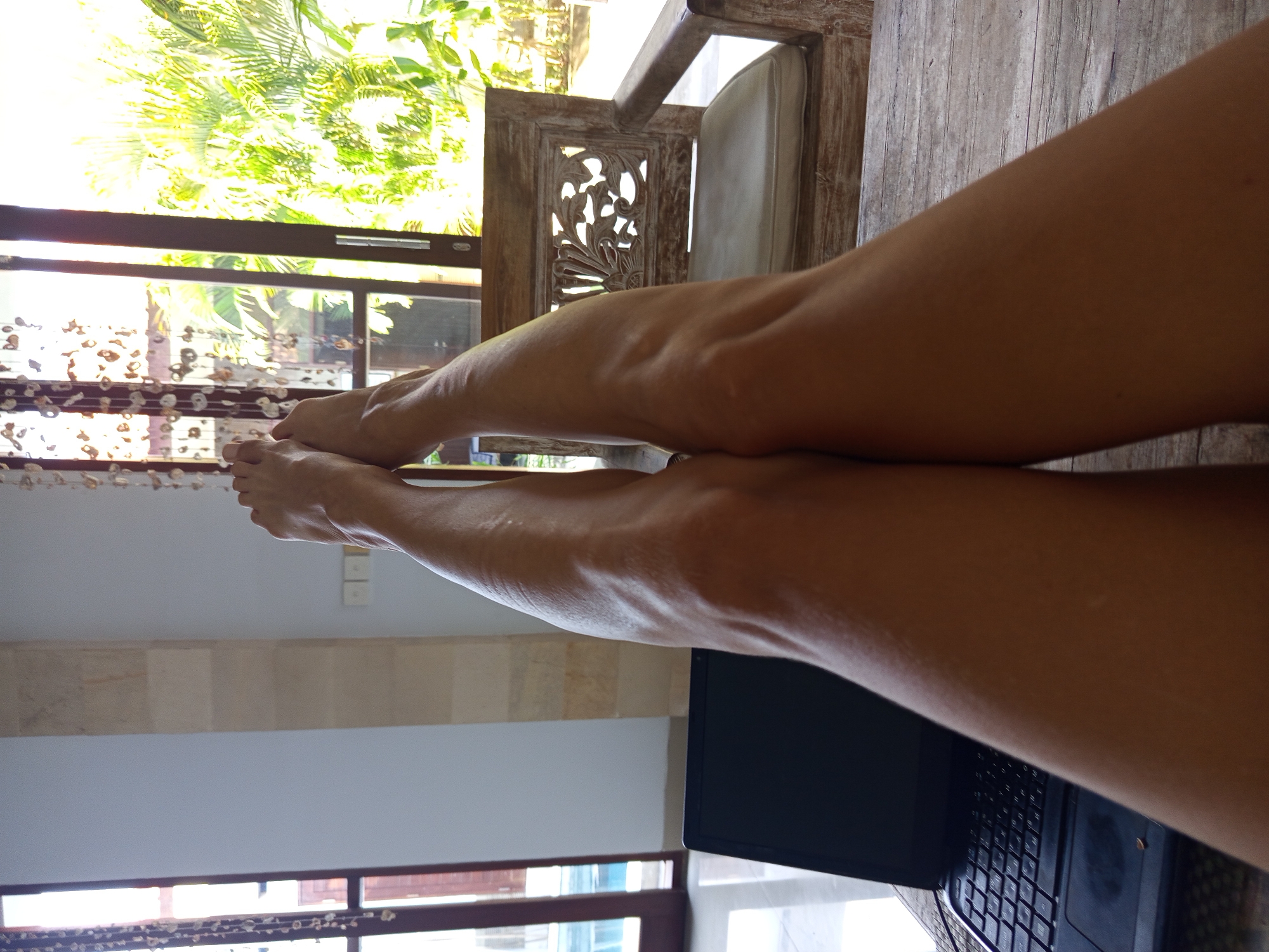 A close-up, indoor shot features a person's legs stretched out, with feet near a window and a laptop visible in the foreground. The setting appears to be a room with an open window offering a view of tropical foliage. The furniture includes a carved wooden chair. Based on the location provided, the scene is likely located in North Kuta, Indonesia. The individual's legs are positioned towards the viewer, implying a relaxed or reclining posture. The laptop suggests a work or leisure activity.