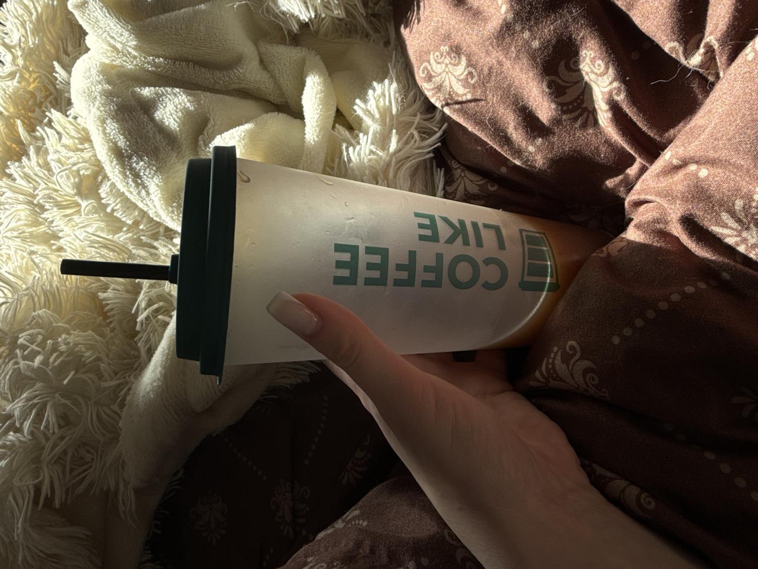 A close-up, top-down view captures a cozy indoor scene, likely a bed or sofa. The primary subject is a person's hand gently holding a frosted, clear tumbler with a dark green lid and straw. The tumbler appears to contain a light-colored beverage, possibly iced coffee, indicated by condensation on its exterior.
The tumbler features text written in a light green color, which reads "COFFEE LIKE" when viewed upright, though it appears upside down from the current perspective. Adjacent to the text is a stylized battery icon, suggesting an energy boost.
The hand, with neatly manicured nails, rests amidst two distinct textures of bedding. To the left, a soft, plush white blanket with long, shaggy fibers creates a fluffy texture. To the right, a brown duvet or blanket with a subtle pattern of lighter brown floral or paisley motifs is visible.
Strong, directional sunlight illuminates parts of the scene, particularly the white blanket and the cup, casting distinct shadows and creating a high-contrast aesthetic. This lighting suggests either early morning or late afternoon. The overall impression is one of comfort, relaxation, and a tranquil moment enjoying a beverage. No specific location cues are visible beyond a comfortable indoor setting.