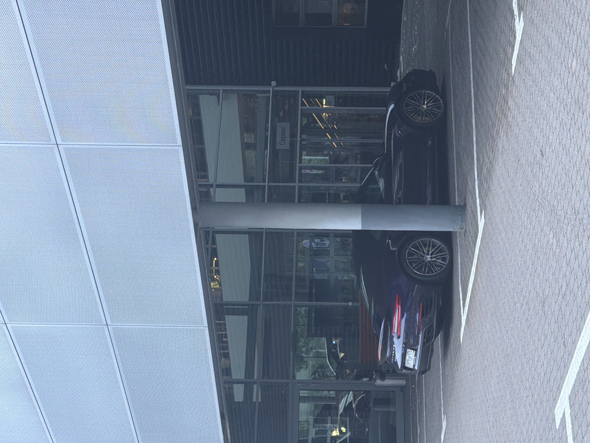 A dark purple Porsche sports car is parked in a lot paved with light gray interlocking bricks. The car is angled towards the right of the frame, with its rear visible. White parking lines are painted on the pavement. Behind the car is a modern building with a perforated silver facade and large glass windows. A white pillar is positioned in front of the car, partially obscuring it. A sign with Russian text, "Сервис," meaning "Service," is visible above the entrance to the building. The overall scene appears to be an outdoor area of a service center or dealership.