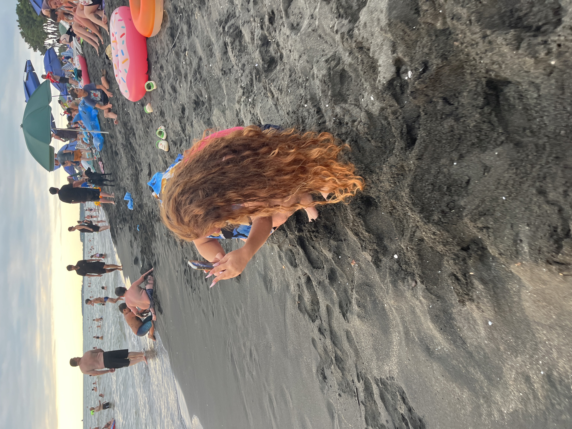 The image depicts a crowded black sand beach in Ureki, Georgia, likely during the day. The foreground is dominated by the dark sand, with footprints and scattered shells visible. A young person with long, auburn, curly hair is kneeling on the sand, holding a smartphone and appearing to take a picture. They are wearing a blue and white patterned top. In the mid-ground, various people are present on the beach, some wading in the shallow water, while others are sitting or standing on the sand. Several beach umbrellas, some green and some blue, are set up, providing shade. Inflatable toys, including a pink donut and an orange ring, are scattered on the sand. Further back, more beachgoers are visible, as well as the sea and a hazy horizon. The overall scene suggests a busy summer day at a popular tourist destination.