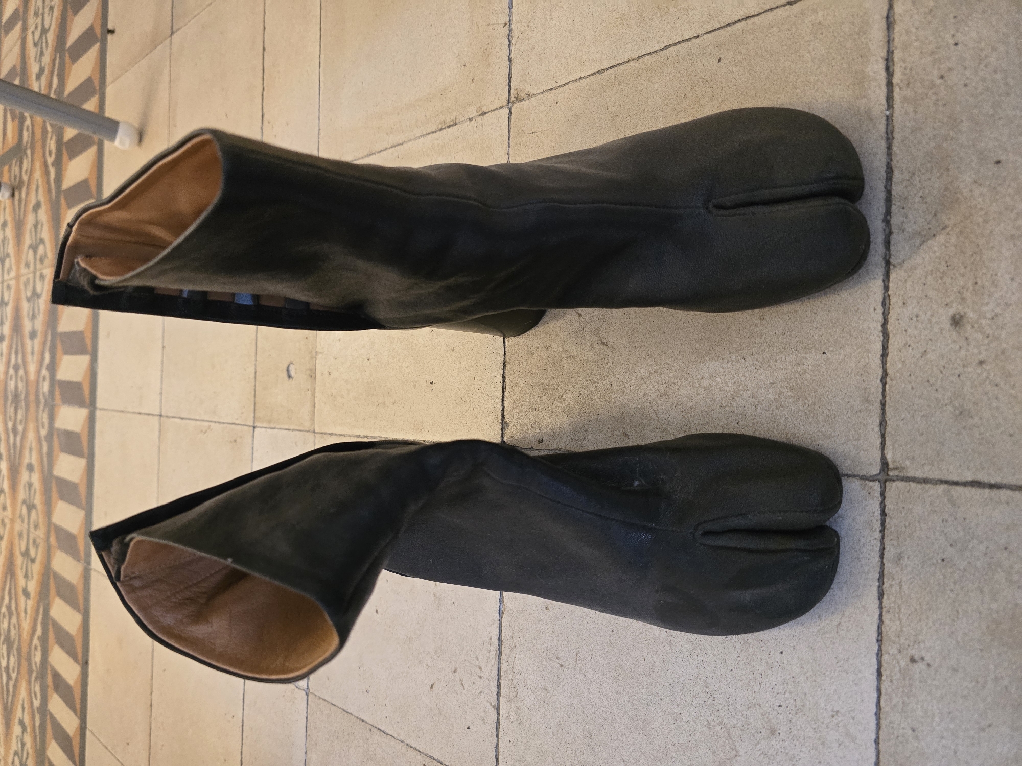 Two black, knee-high boots with a distinctive split-toe (tabi) design are laid side-by-side on a tiled floor, viewed from an overhead perspective. The boots appear to be made of leather, showing some creasing and slight wear, suggesting a soft, flexible material. The interior lining visible at the top opening of each boot is a lighter, tan or brownish color.
The floor beneath the boots consists primarily of light beige or off-white square tiles with dark grey grout lines. To the left side of the frame, a section of the floor features an ornate, geometric pattern with brown, orange, and darker-toned tiles, creating a decorative border or a different flooring zone.
No people are visible in the image, and the boots simply rest on the floor, perhaps indicating storage, display, or a pause in wear. There are no discernible weather conditions, specific time of day cues, or emotional expressions. No text is visible anywhere in the image. The setting appears to be indoors, possibly within a home or commercial space.