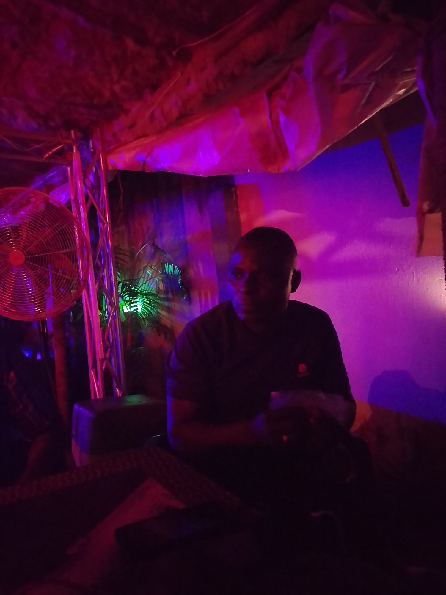 A dark-skinned male is seated in a dim, purple and red-lit environment, possibly indoors. The scene includes a fan, stage lighting equipment, and what appears to be foliage in the background. The location is Unknown, Unknown. The subject is looking off-camera. The foreground is obscured.
