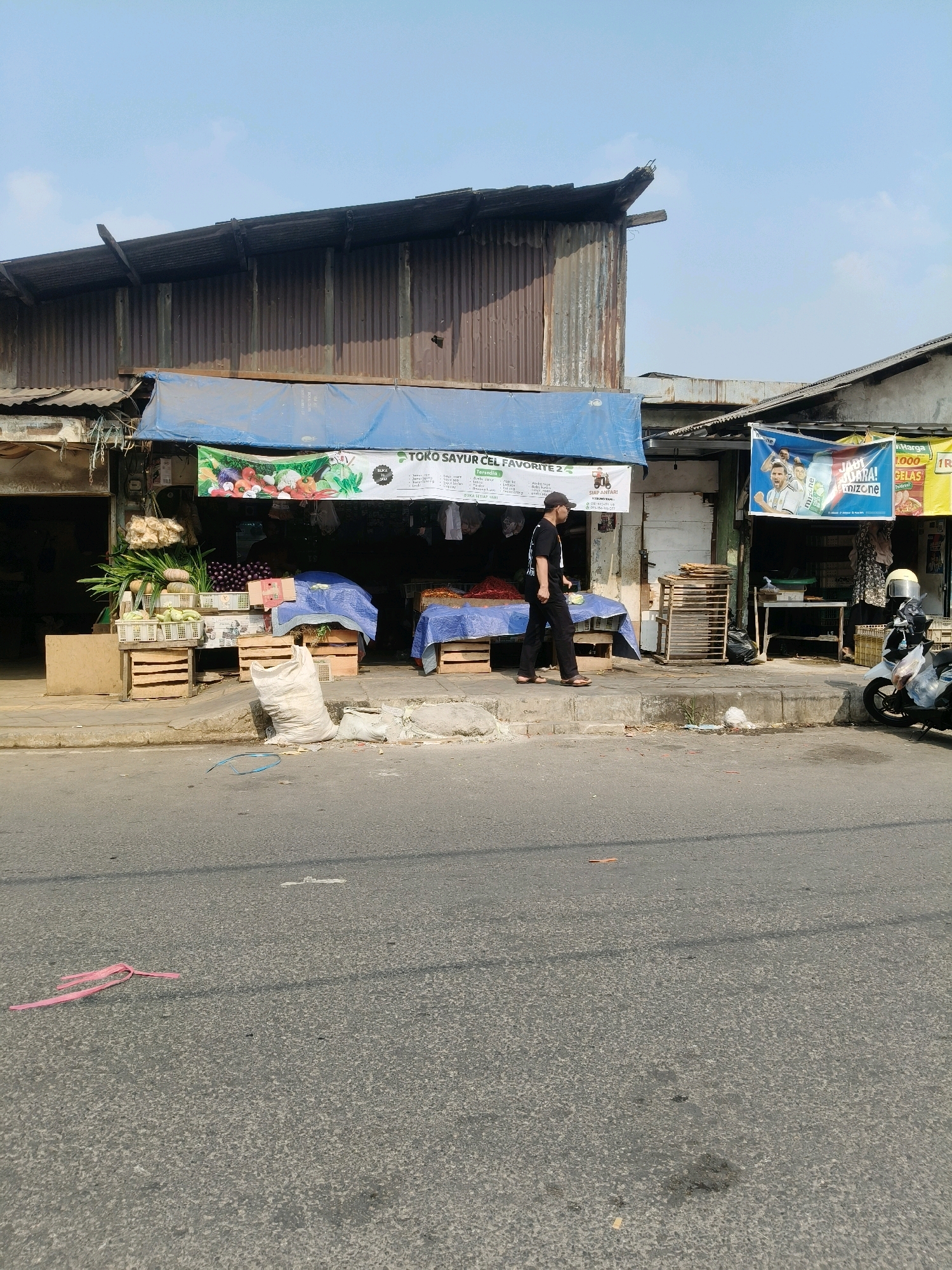 This is a medium shot, eye-level, outdoor photograph taken on a sunny day in the city of South Tangerang, Indonesia, likely on a street or roadside market. The scene depicts a row of small market stalls, characterized by corrugated metal roofs and simple wooden structures. In the foreground, a paved road is visible with some debris, including a pink plastic strap. A concrete sidewalk runs alongside the road, where several market stalls are set up.
One stall prominently displays fresh produce, with crates of vegetables like cabbage and leafy greens. Another stall has a blue tarp covering its wares. A man wearing a black t-shirt, black pants, and a black cap is walking on the sidewalk from left to right, away from the viewer. He is carrying a white bag in his left hand. Behind the stalls, various goods are displayed, including what appears to be more produce and stacked wooden crates. Several banners are hung above the stalls. One banner, partially obscured, shows an image of vegetables and text including "TOKO SAYUR CEL FAVORITE 2". Another banner features an image of a soccer player, likely Lionel Messi, with text in Indonesian. A white scooter with a helmet on its seat is parked on the sidewalk near the right edge of the frame. The environment appears to be a bustling commercial area, typical of a roadside market in Indonesia.
