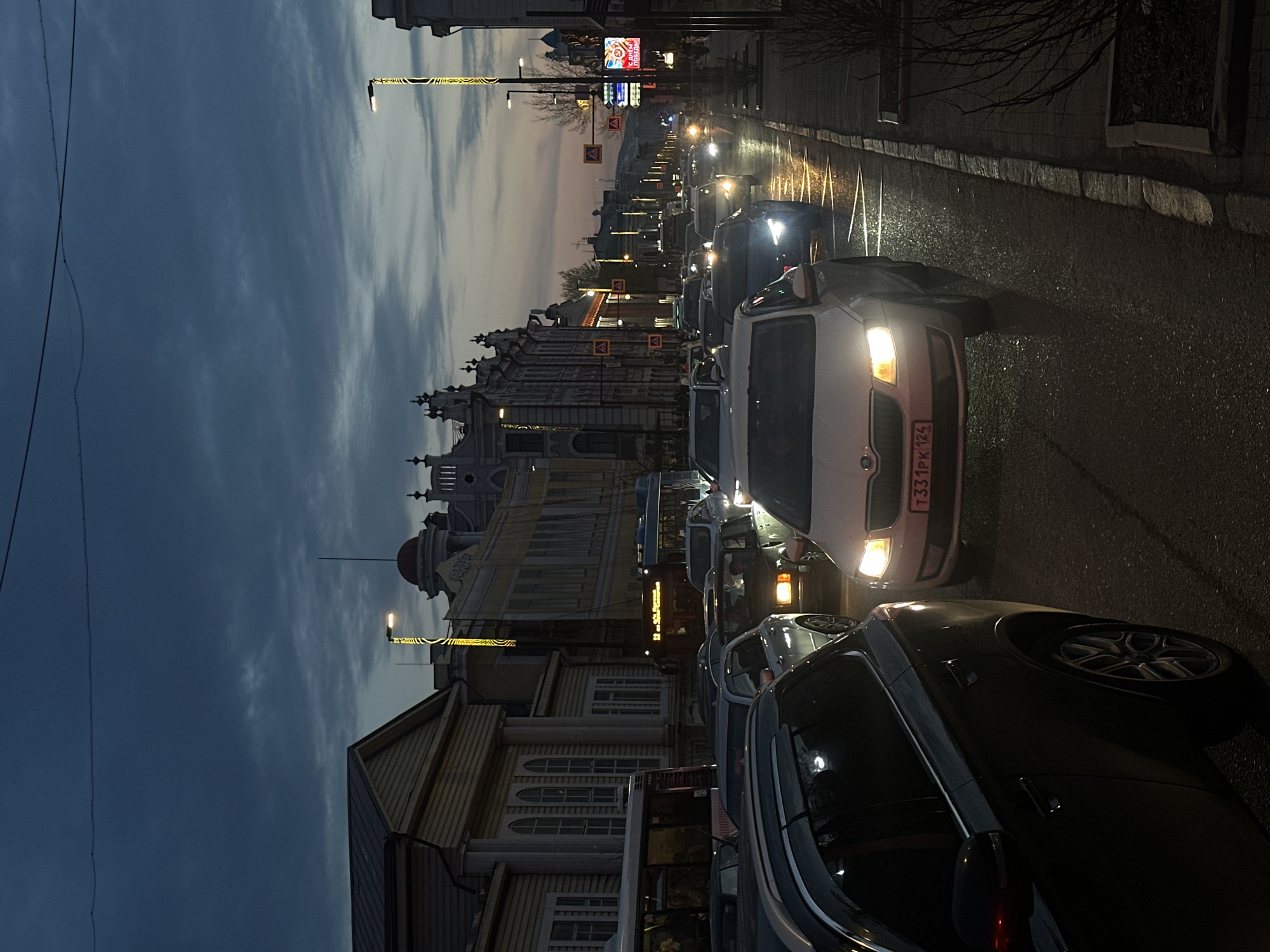 The image shows a street scene in Krasnoyarsk, Russia at dusk. Several cars are driving on a wet road, with the lights of the vehicles reflecting on the pavement. Buildings line the street, including a historic building with ornate architecture and a wooden structure with arched windows. Streetlights illuminate the scene. The sky is cloudy.