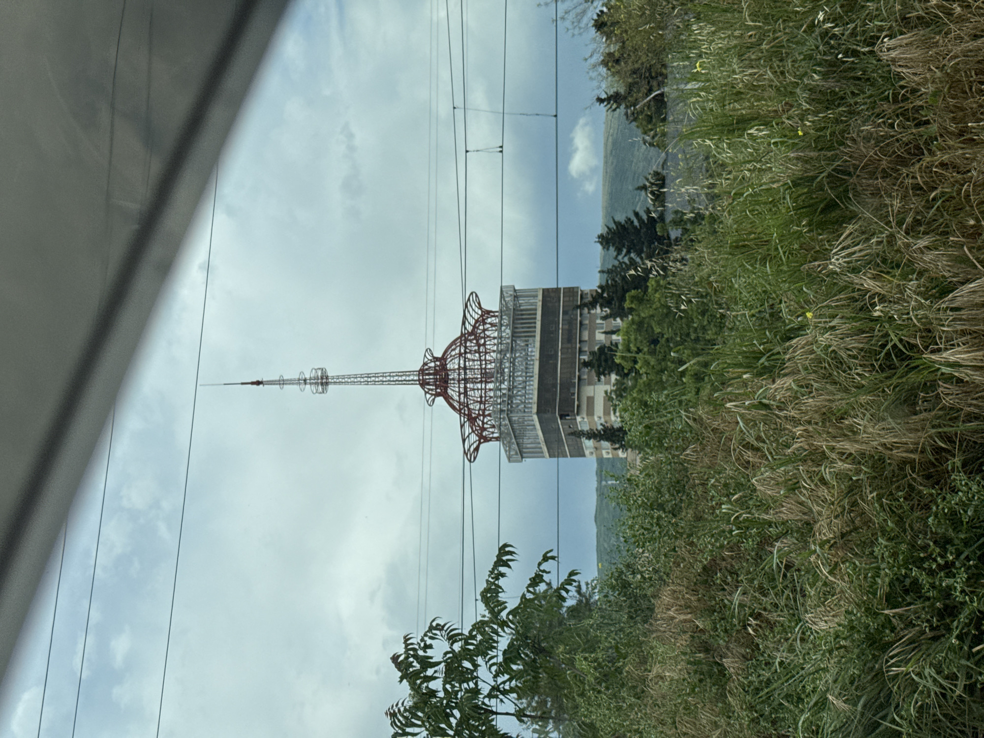 The image captures a view of the Tbilisi TV Tower, a prominent landmark in Tbilisi, Georgia. The tower, with its distinctive lattice structure and antenna, rises above a dense collection of trees and vegetation in the foreground. The sky is overcast with clouds, and power lines are visible crisscrossing the upper portion of the frame. The lower part of the tower appears to be a rectangular building with several stories. The scene is likely captured from a distance, possibly from a vehicle given the slight blur and the perspective.