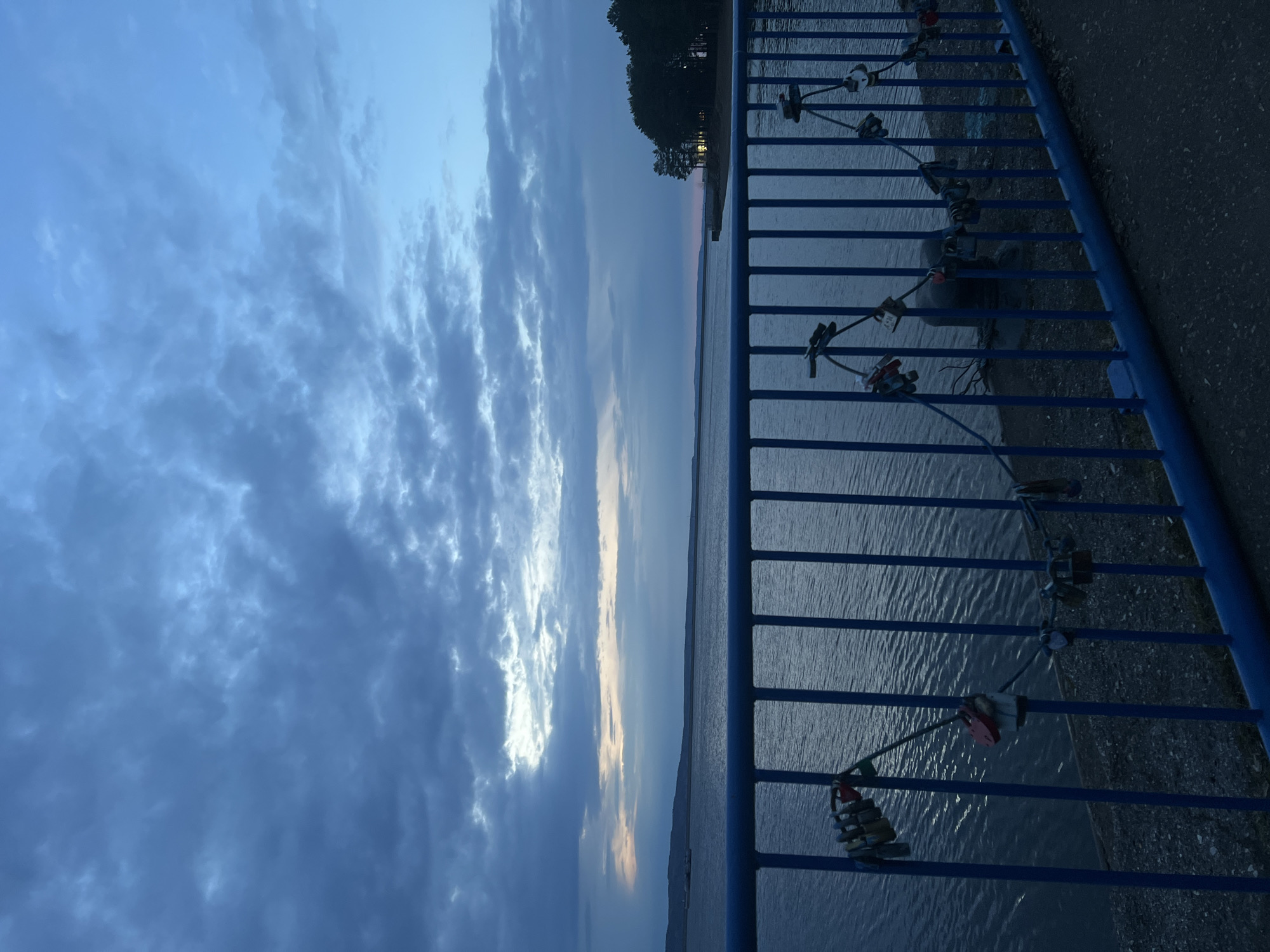 This image captures a serene view over a vast body of water, likely the Votkinsk Reservoir in Chaikovsky, Russia, at either dawn or dusk. The scene is dominated by a clear blue sky filled with varied cloud formations, transitioning from deep blues at the top to lighter, softer hues of orange and pink near the distant horizon.
In the foreground, a sturdy blue metal railing runs diagonally across the right side of the frame, serving as a boundary for a paved pathway that borders the water. Attached to the vertical bars of this railing are numerous "love locks" of different shapes, sizes, and colors, including red and silver padlocks. These locks signify a common romantic tradition.
Beyond the railing, the calm, dark blue surface of the water extends towards the horizon. The water shows subtle ripples and reflections of the sky's light. A distant shoreline, appearing as a dark silhouette of trees, is visible across the water, with a faint glow from the setting or rising sun illuminating the sky directly above it.
The overall atmosphere is tranquil and contemplative, with the soft, dim light characteristic of the "golden hour." No people are visible, and while some locks might have engravings, no specific text is discernible. The image evokes a sense of peace and enduring sentiment.