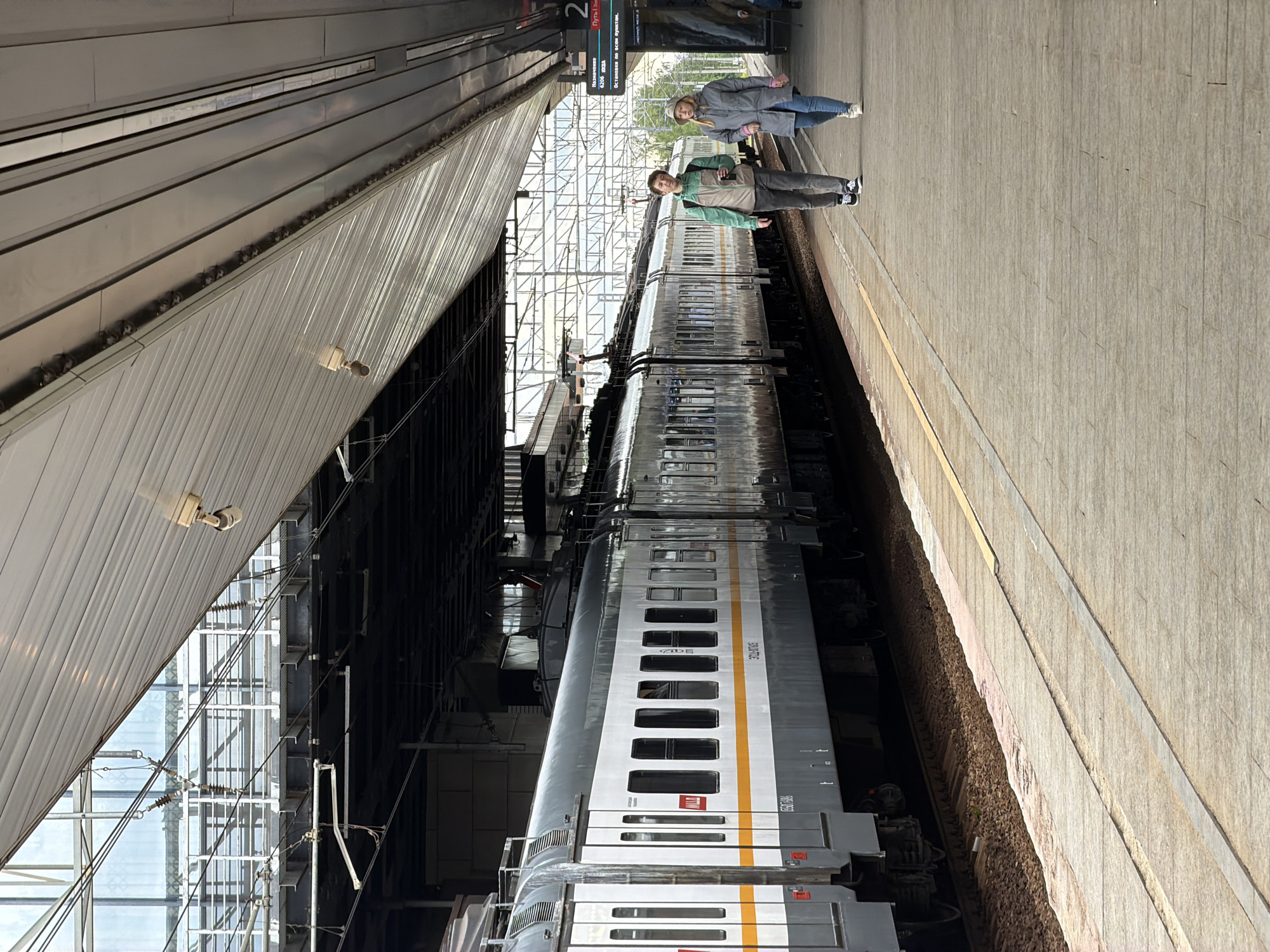 The image depicts a train station platform in Moscow, Russia. A silver and gray train is stationary at the platform. Two individuals, a male and a female, stand on the platform near the train. Signage indicating "Путь 2" (Track 2) is visible overhead. The platform is constructed of light gray material. The station has a high ceiling with both solid and transparent panels. Overhead electrical wires and structural supports are prominent.