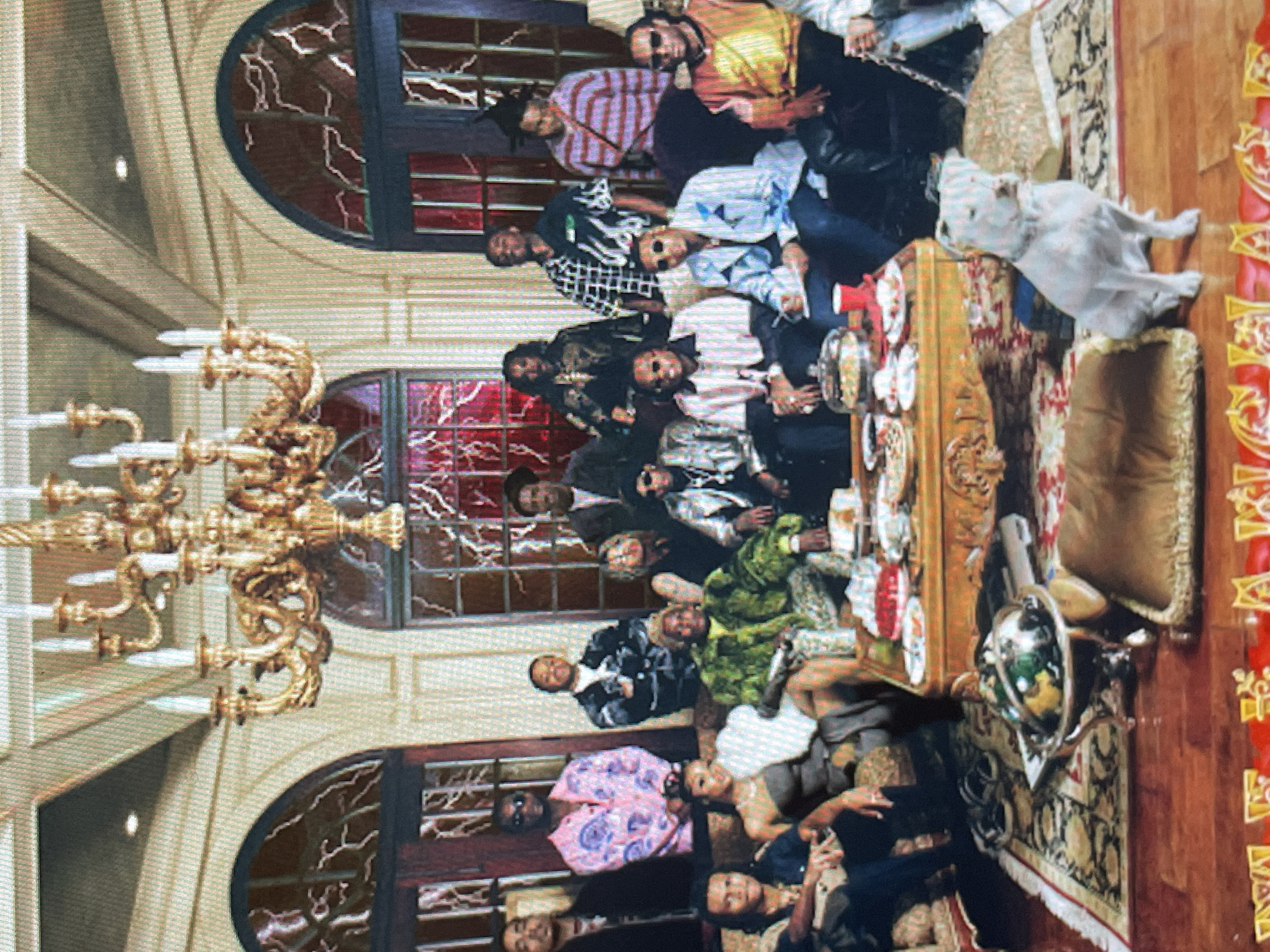This media file captures a large group of people, predominantly young adult males, gathered in a luxurious, ornately decorated room. The image is rotated 90 degrees counter-clockwise.
**Subjects:** Approximately 15-20 individuals are visible, mostly men, dressed in fashionable casual attire. Many are wearing sunglasses, and their expressions range from relaxed to confident. The room features an elaborate, multi-branched golden chandelier hanging from a high ceiling, though its lights are unlit. Large arched windows dominate the background, featuring distinctive red, crack-like patterns in the glass, giving them an almost stained-glass or fractured appearance. An ornate wooden table, laden with dishes and glasses, is central to the gathering. Other objects include patterned rugs, plush seating, a white statue of a dog (resembling a bull terrier), and a globe on a stand.
**Activity or event taking place:** The scene depicts a social gathering or party, with the individuals posing together for a photograph. They are standing, sitting, and leaning around the table and throughout the room, suggesting a casual yet celebratory atmosphere.
**Notable details:** The setting is opulent, characterized by the grand chandelier, intricate architectural details on the walls and ceilings, and rich furnishings. The red patterns within the windows create a dramatic and somewhat mysterious backdrop, obscuring any view of the outside and making the time of day undeterminable. The overall aesthetic suggests a high-end, private event in a stately venue. The location context provided is Sofia, Bulgaria.
**Visible text:** No decipherable text is visible anywhere in the image.