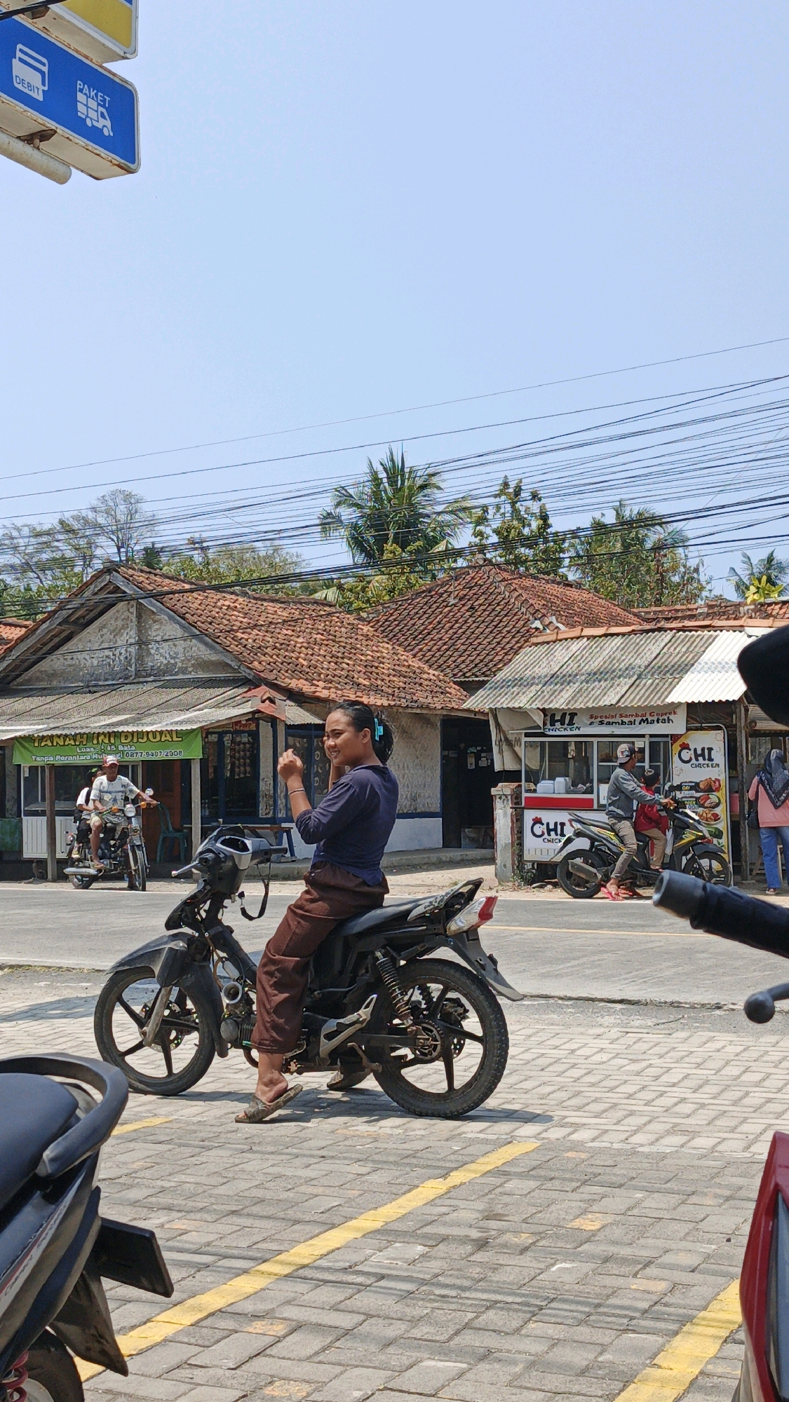 The image shows a street scene in Cibenda, Indonesia, under a clear blue sky. In the foreground, a woman is riding a black motorcycle. She has her hair pulled back and is wearing a dark blue long-sleeved shirt and brown pants. She appears to be looking to her right and holding something in her hands near her face. The motorcycle has a license plate. Behind her, another person is visible on a motorcycle, and further back, there are several buildings with tiled roofs. One building has a green banner that reads "TANAH INI DIJUAL" (This land is for sale). To the right of that, there is a food stall with signs that say "CHI CHICKEN" and "Sambal Matak". Other motorcycles and people are also visible in the midground and background, suggesting a moderately busy street. Overhead, power lines are strung across the sky. The street is paved with interlocking concrete blocks, and yellow lines mark a pedestrian crossing.