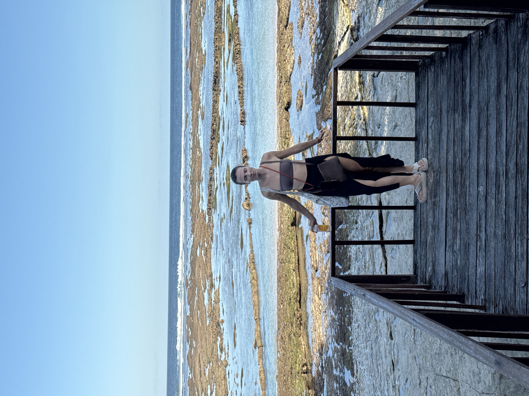 A woman stands on a wooden deck or boardwalk in Kuta Selatan, Indonesia. She has a light brown bandeau-style top, a black skirt, a cross-body bag, and white sandals. She holds a yellow object, possibly a drink, in her left hand. The deck overlooks a shallow ocean area with exposed rock formations and tide pools during low tide. The sky is blue.