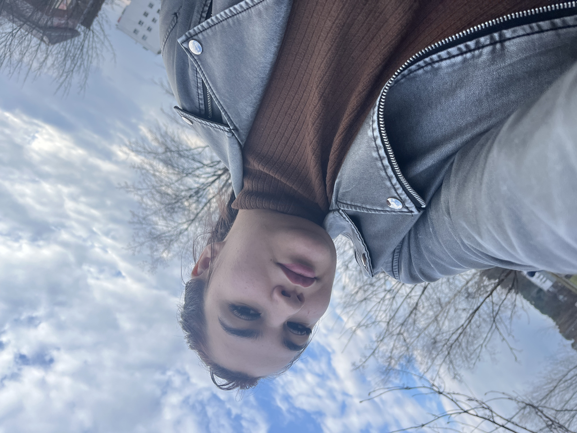 A young woman is taking a selfie outdoors in Chaikovsky, Russia. She is wearing a brown turtleneck sweater and a gray leather jacket. The background includes a cloudy sky, bare trees, and buildings. The orientation of the image is inverted, as if she is looking down at the camera.