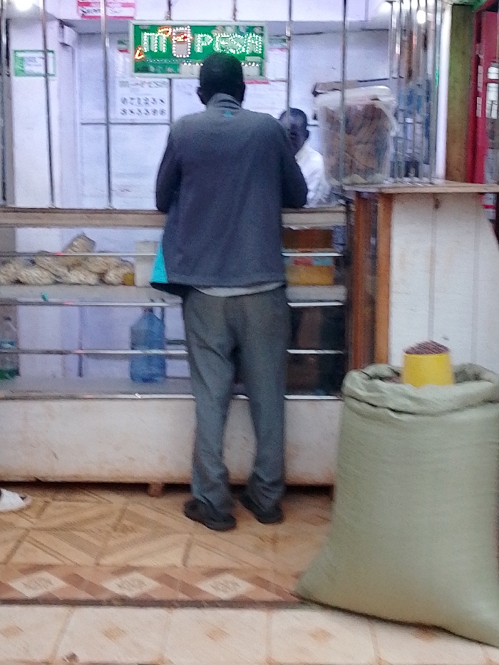The image shows a man standing in front of a counter inside a small shop in Kivaywa, Kenya. The man is wearing a blue jacket and gray pants. He is looking down at a counter that has a large green bag sitting on the floor in front of it. Behind the counter are shelves with various objects including a bottle of water. The counter is made of wood and has a white painted top. The floor is tiled with brown and beige tiles. The shop's walls are white and have a sign that reads "M-PESA." The location based on the coordinates is a small shop on a street in Kivaywa, Kenya. The image is taken from a slightly low angle, giving the viewer a perspective from the man's point of view. The photo is in focus and the lighting is dim. The image was taken on a sunny day.