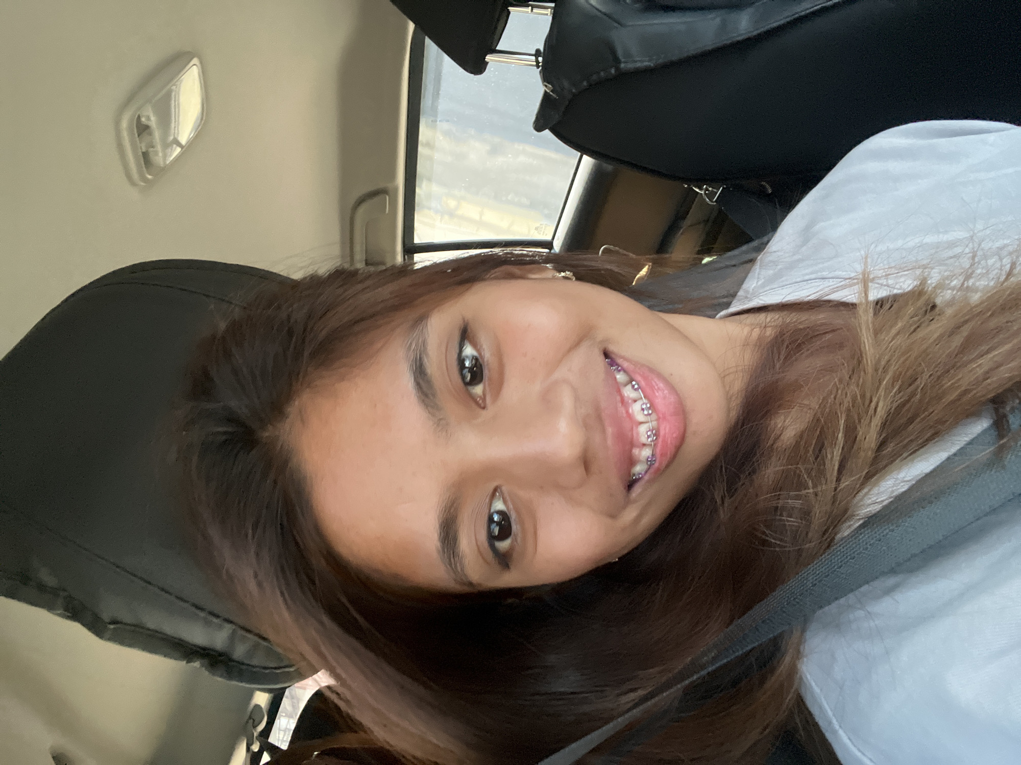 A young woman, appearing to be of Southeast Asian descent, is depicted from the upper chest upward, seated in the front passenger seat of a vehicle in Davao City, Philippines. She has long, dark brown hair, dark brown eyes, and is smiling, revealing dental braces with pink or purple elastic bands on her upper teeth. She wears a light-colored (white or light grey) t-shirt and a grey fabric seatbelt across her left shoulder and chest. Small, silver-colored hoop earrings are visible. The car's interior shows a light-colored ceiling with a rectangular overhead light fixture, and a black headrest with two silver metal posts is visible to her right. Through the car window on the right, a blurred outdoor background is visible, suggesting daylight conditions.