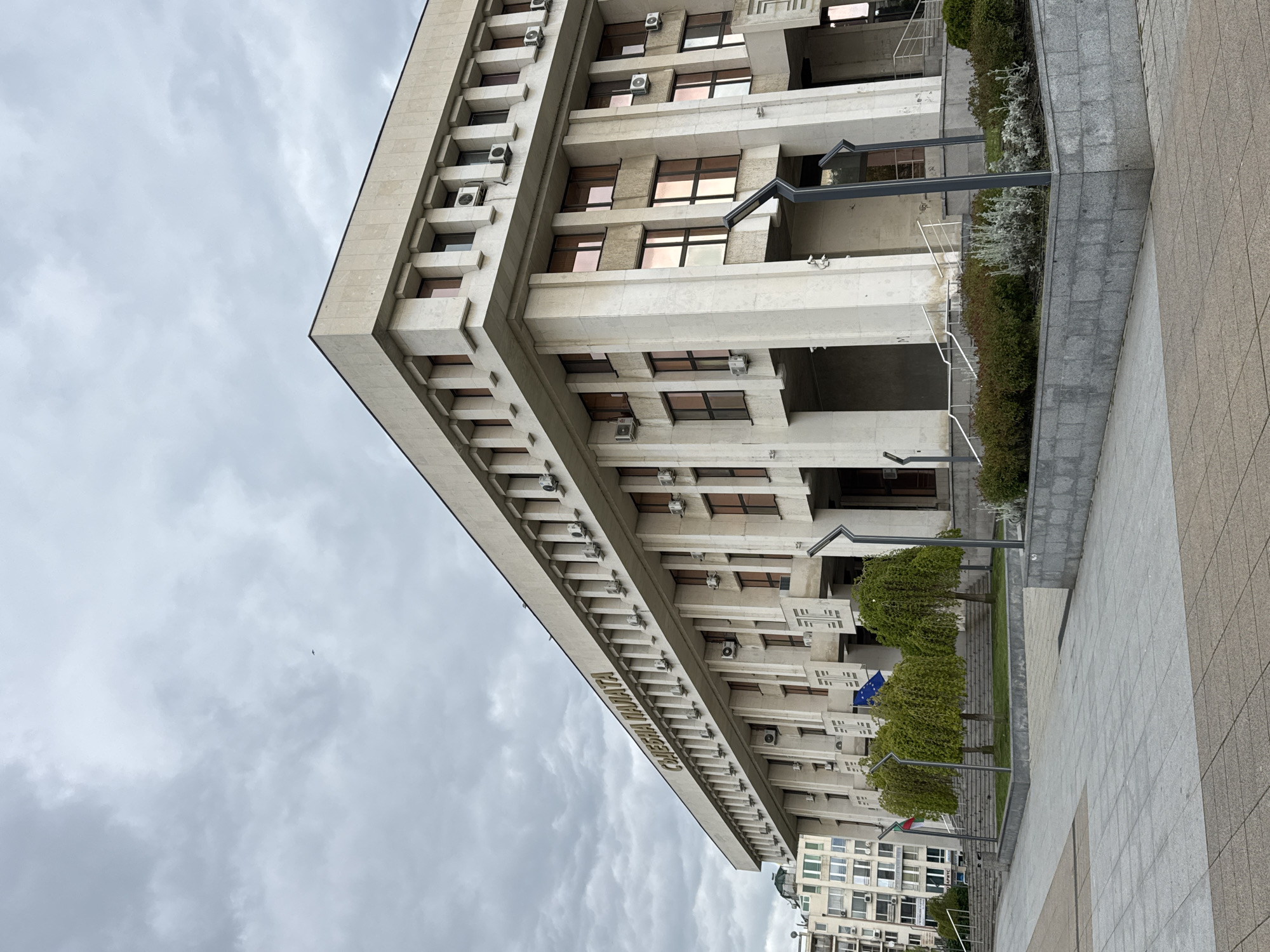 The image depicts a low-angle view of a multi-story building, identified by the inscription "СЪДЕБНА ПАЛАТА" (Courthouse) in Cyrillic on its facade, suggesting its purpose. The architecture is characterized by a repetitive pattern of windows, horizontal bands, and numerous air conditioning units attached to the exterior walls.
The building is located in Burgas, Bulgaria, as evidenced by the Bulgarian and European Union flags displayed near the base. The surrounding environment includes paved pedestrian areas, landscaping with low shrubs and manicured trees, and street lighting. The sky is overcast with gray clouds. There is a hint of other buildings in the far background. There are no people visible in the image.