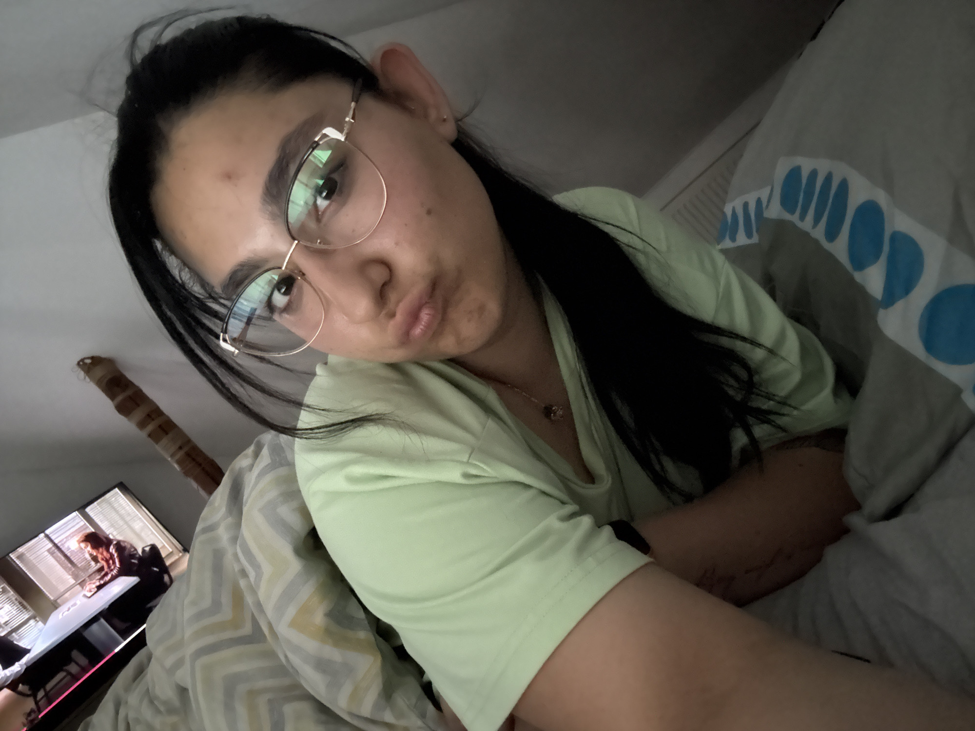 A person with dark hair pulled back, wearing round eyeglasses with gold frames and a light green t-shirt, is lying in bed. The person is looking at the camera with pursed lips. The bed has patterned bedding in shades of gray, white, and yellow and another patterned blanket in light gray and blue. In the background is a television showing a person sitting at a desk. The location is likely indoors, possibly a bedroom, and the city of Elhovo, Bulgaria, is not apparent from the visual details.