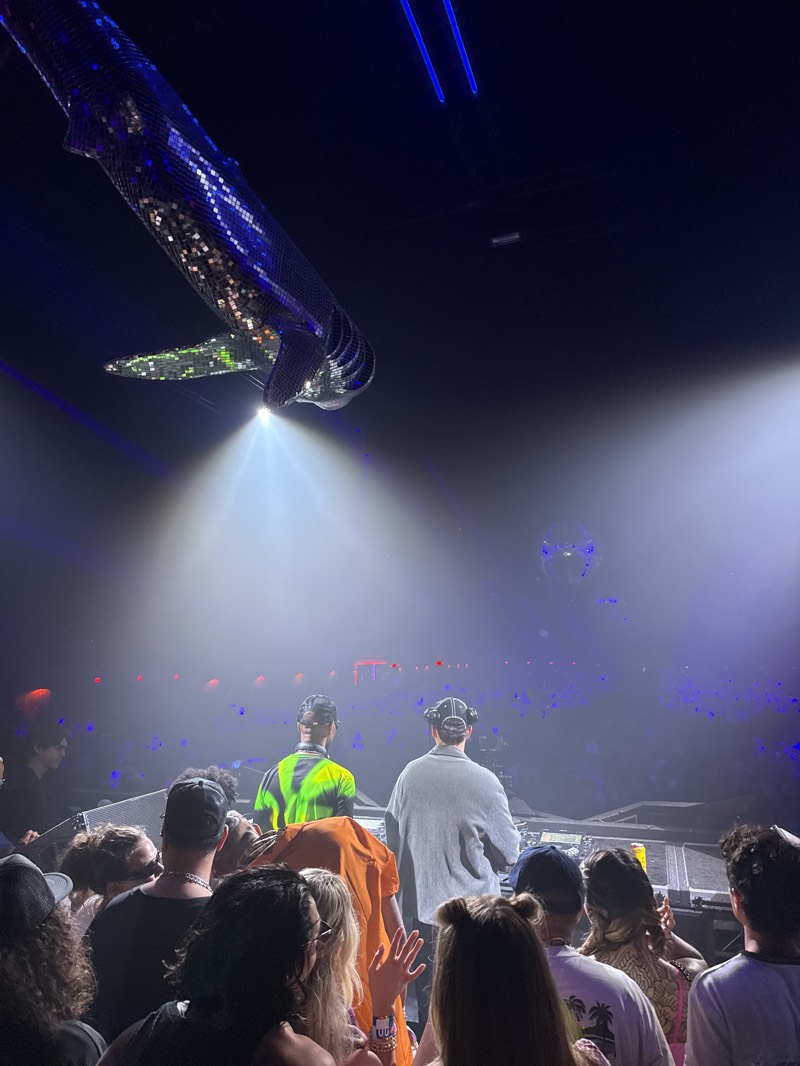 The image depicts a crowd of people at a concert venue in Indio, United States, at night. Two figures, presumably performers or DJs, stand on a platform facing the audience, illuminated by bright spotlights. The crowd is dense, with many faces visible. Overhead, a large, reflective object shaped like a whale hangs from the ceiling, emitting light. The atmosphere is dark with some blue and purple lighting.