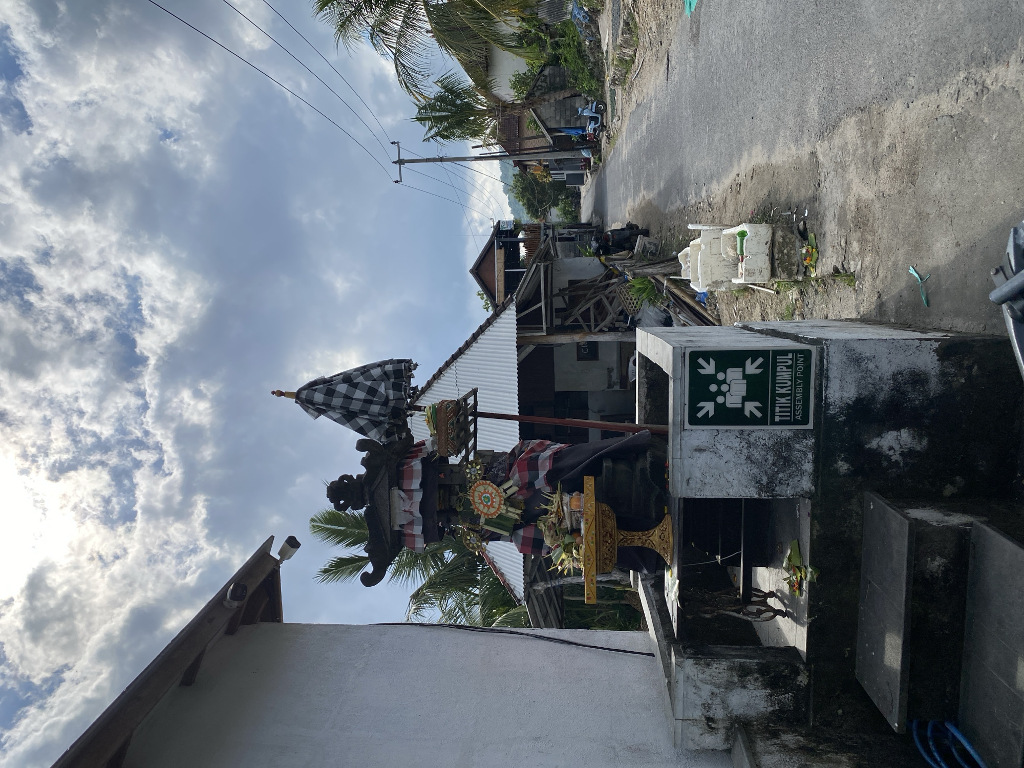 This media file captures a scene in Nusa Penida, Indonesia. The primary subject is a traditional Balinese statue, adorned with a black and white checkered umbrella, sarongs in red and white, and decorative offerings. The statue is positioned in front of a building with a corrugated metal roof and appears to be part of a local shrine or decoration.
Behind the statue, a paved road runs through a village. The setting includes residential structures, some appearing under construction or in disrepair, with visible wooden beams and scaffolding. Tropical vegetation, including palm trees, is present throughout the scene, indicating a warm climate. Electrical wires are strung across utility poles, a common sight in many Indonesian villages.
To the right of the statue, a concrete structure displays a green and white sign with text that reads "TITIK KUMPUL ASSEMBLY POINT". The sign also features an icon with arrows pointing towards a group of people, suggesting a designated meeting or evacuation point. Nearby, there are white plastic containers and some miscellaneous debris, hinting at ongoing activity or a less-than-pristine environment. A scooter is parked further down the road.
The sky is partly cloudy, with patches of bright sun breaking through, suggesting it is daytime, likely late morning or early afternoon. The overall atmosphere appears calm and possibly humid, typical of a tropical locale. No people are directly visible interacting with the statue or the immediate surroundings, lending a sense of quietude to the scene. The composition is a vertical orientation.