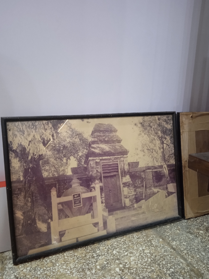 The image shows a framed black and white photograph of a stone building with a wooden door. The building appears to be a temple or shrine, with a tiered roof and stone walls. The photograph is displayed on a wall in a room with a tiled floor. The wall is white with a light grey tint, and the tiled floor is grey with white speckles. There is a second framed picture to the right of the first. The first picture is mounted in a black frame, while the second is mounted in a wooden frame with a light brown hue. The second picture has a white background and an indeterminate subject matter. There are no people or objects in the room besides the two framed pictures, which are against the wall. The image is taken from a perspective looking directly at the pictures. The photograph was likely taken in Surakarta, Indonesia, at the geographic coordinates of -7.559 latitude and 110.783 longitude.