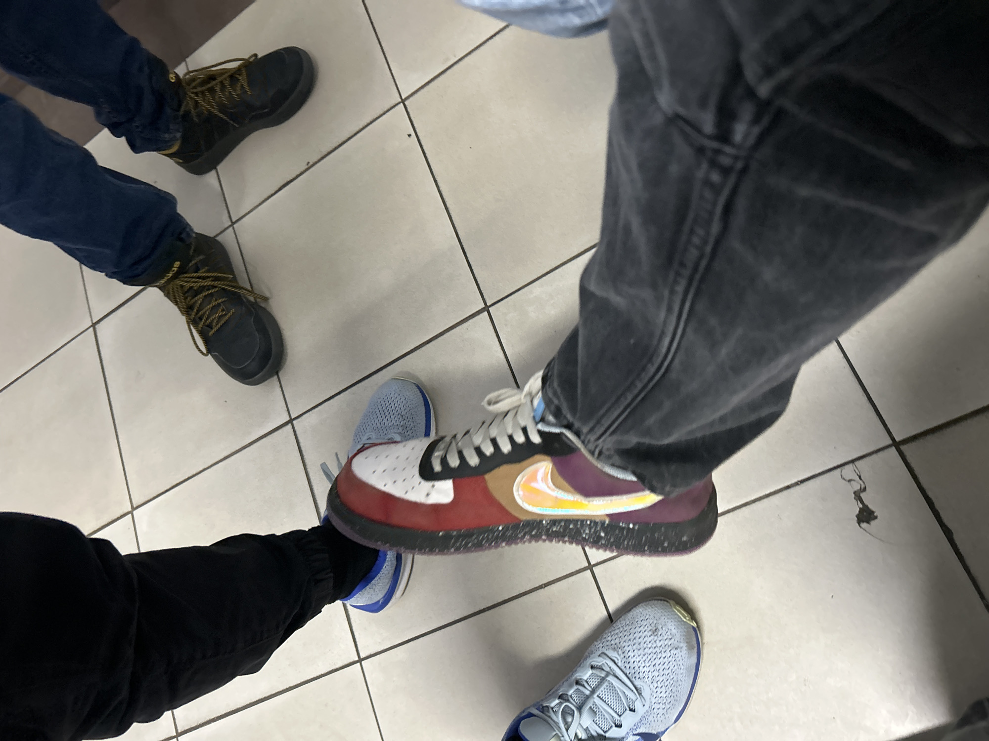 The image depicts four individuals standing on a tiled floor. Two individuals on the left wear dark-colored boots and dark blue pants. A third individual is wearing black sweatpants and light blue sneakers. The fourth individual wears gray jeans and stands on the third individual's foot while wearing a multi-colored sneaker. The tiled floor consists of light-colored square tiles. The specific location in Burgas, Bulgaria, cannot be determined from this image.