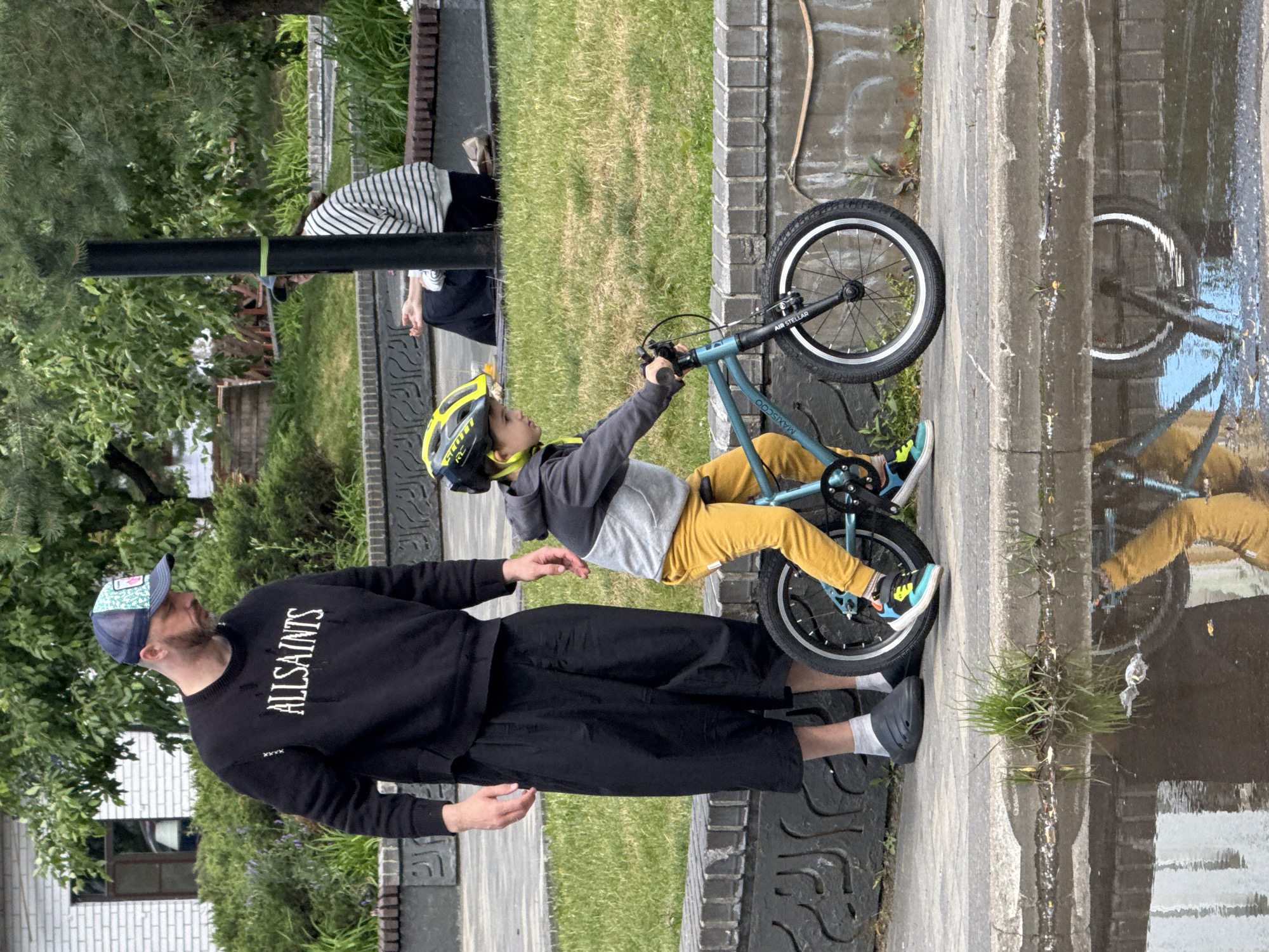 In Moscow, Russia, a father stands beside his young son who is on a bicycle. The father, wearing a black sweatshirt with "ALLSAINTS" printed on it and a baseball cap, is looking down. His son, sporting a yellow helmet, is seated on a teal bicycle with the name "MAXISCOO" visible on the frame. The boy is wearing yellow pants and colorful sneakers. They are on a paved sidewalk next to a grassy area with trees and a brick planter. In the background, another person is sitting on the grass. A puddle of water reflects the scene and the sky.