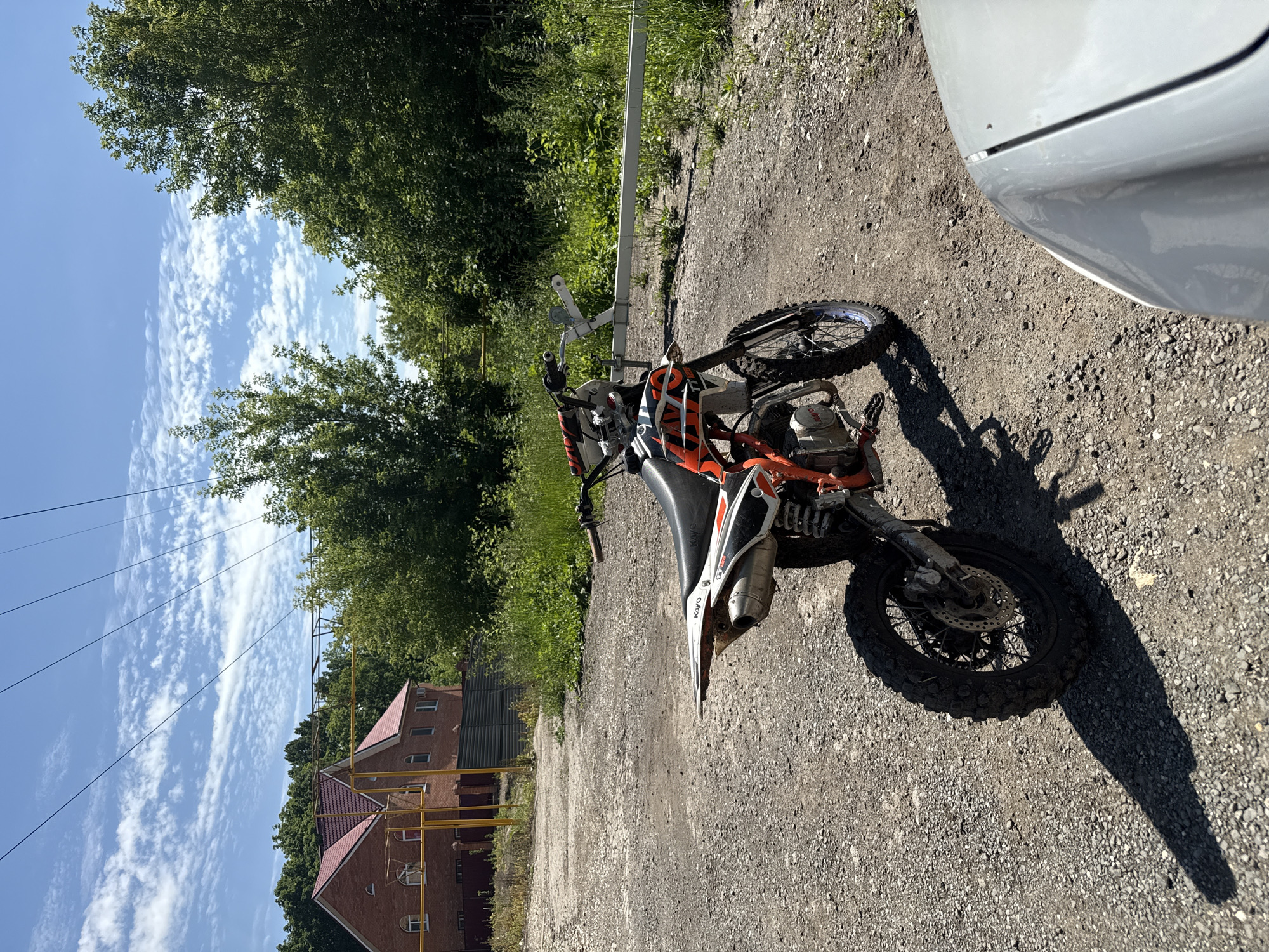 An orange and black dirt bike is parked on a gravel surface in front of a white car. In the background, a brick house with a red roof and a metal fence are visible. Trees and vegetation are abundant, and power lines are strung across the bright blue sky, which is dotted with white clouds. The scene appears to be an outdoor location, possibly in a suburban or rural area. There are no people visible in the image.