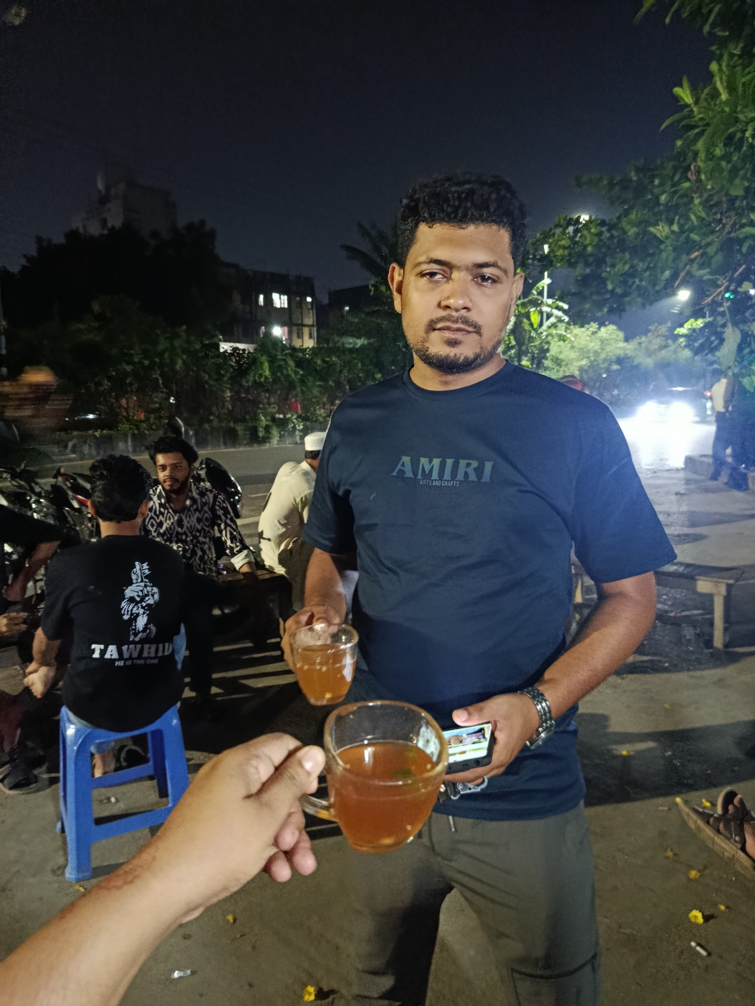 The image captures a nighttime scene in Dhaka, Bangladesh. A man wearing a dark blue AMIRI t-shirt and grey pants is the central figure, holding a cup of tea. Another hand reaches into the frame, offering a second cup. The man also holds a phone in his other hand and is wearing a silver bracelet. In the background, several other men are seated on benches and stools alongside parked motorcycles, suggesting a roadside tea stall or gathering spot. One man wears a black T-shirt emblazoned with "TAWHID", another is wearing a shirt with a patterned design, and a third wears a white cap. Buildings are visible in the distance under a dark sky. The ground is littered with debris. A car is passing on a wet road in the background. Streetlights illuminate the scene, with trees and greenery adding to the urban backdrop.