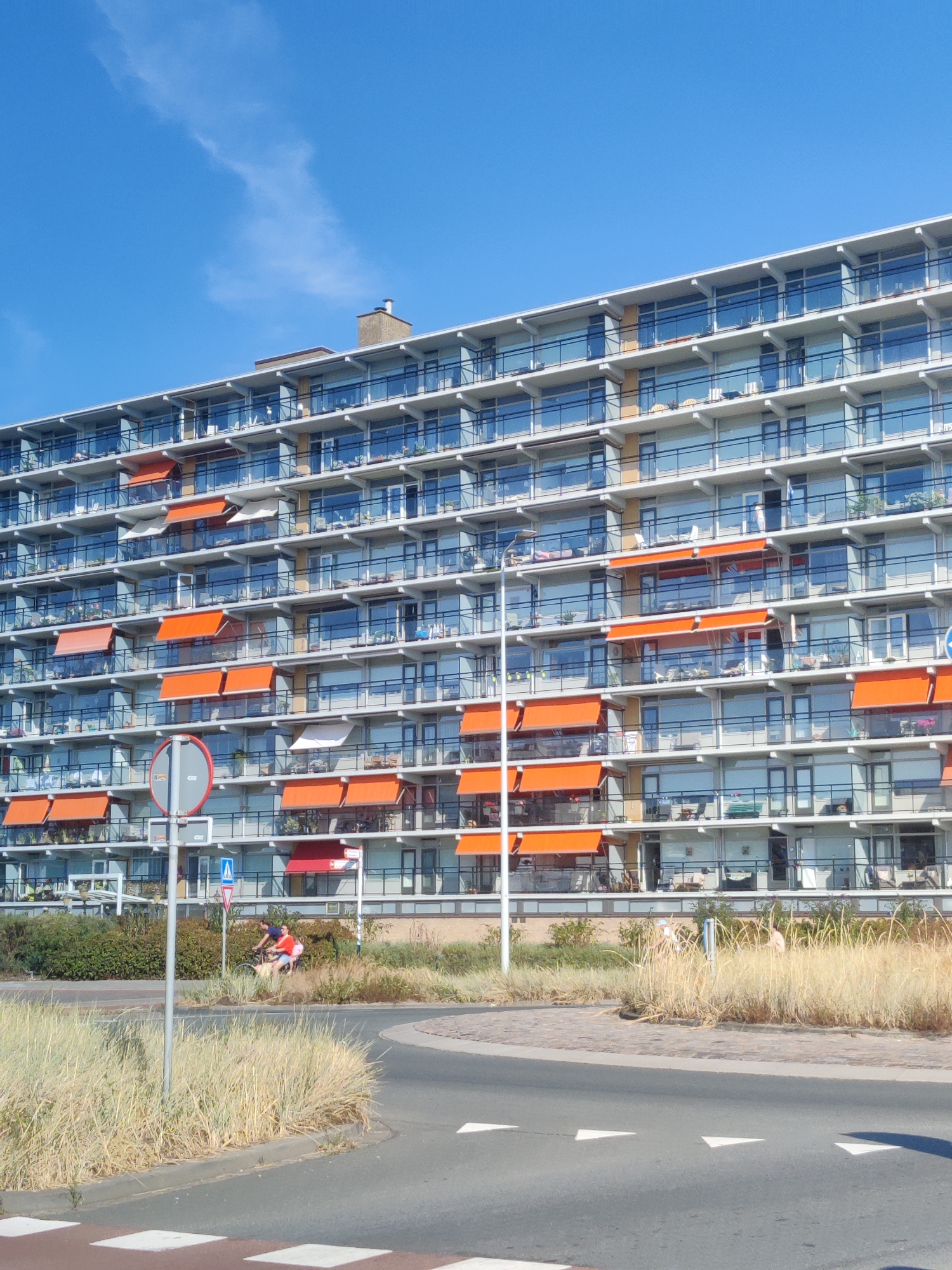 The image shows a large, multi-story apartment building with many balconies and windows. Several balconies have orange awnings extended. The building appears to be located in The Hague, Netherlands, as suggested by the presence of what seems to be a street sign with a Dutch coat of arms. In the foreground, there is a road with white lane markings, and to the left of the road, there is a large signpost with a circular sign. Behind the apartment building, there is an area with tall, dry grass and some greenery. In the middle ground, two people are riding a bicycle through the tall grass, with one person sitting on the handlebars and the other pedaling. The sky is clear and blue.