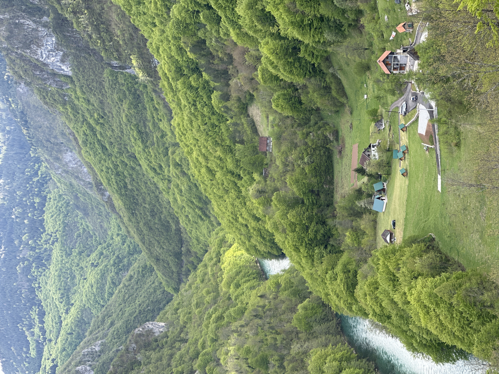 The image depicts a vertical view of a landscape in Đurđevića Tara, Montenegro. A river winds through a valley, flanked by densely forested hills on both sides. The trees are primarily green, indicating a verdant environment. On the right side of the image, there's a grassy area with a cluster of buildings, including small cabins with green roofs and a larger house with a red-tiled roof. A road or pathway connects the buildings. A white car is parked in front of one of the buildings. A rocky cliff is visible on the left side of the image, adding to the mountainous terrain. The overall scene suggests a rural or tourist area within a scenic natural environment.