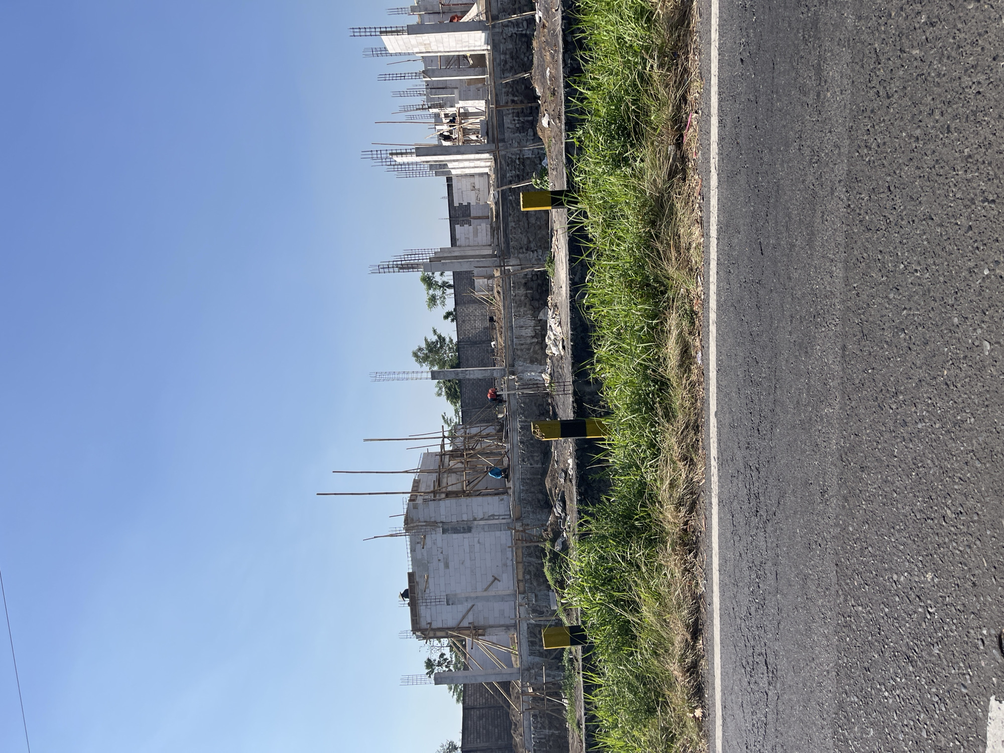 The image captures a construction site in the city of Purworejo, Indonesia, under a clear blue sky. The foreground is dominated by a paved road with a white line marking its edge, and beyond that, a strip of overgrown green grass. In the mid-ground, the construction site is visible, featuring the skeletal framework of a building. Numerous concrete pillars with rebar protruding upwards indicate the ongoing construction of multiple levels. White brick walls are being erected, and scaffolding made of bamboo poles and wooden planks supports the structure. Several workers are present at the site, engaged in construction activities. Two yellow and black striped posts stand along the edge of the construction area, possibly serving as markers. In the background, a grey brick wall encloses the construction site, with some trees and vegetation visible beyond it. The overall scene depicts an active construction project in an outdoor, sunny environment.