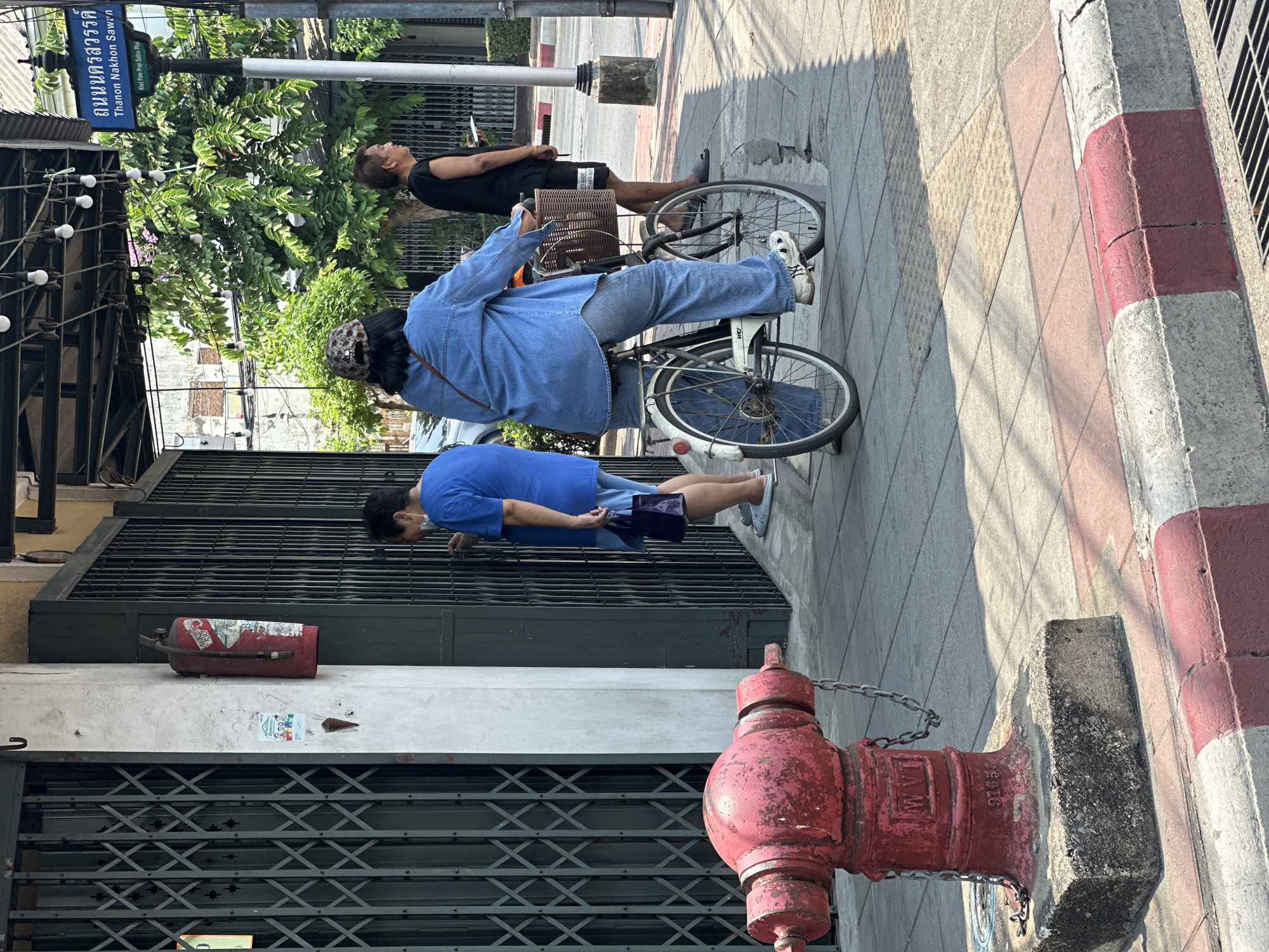 The image shows a street scene in Bangkok, Thailand. Three people are visible on the sidewalk. A man in a black shirt is standing looking up towards the street sign “Thanon Nakhon Sawan”. A woman in a blue denim shirt is standing next to a bicycle. Another person in a blue shirt is standing next to a metal gate. The sidewalk is gray and red-and-white striped. A red fire hydrant stands near the metal gate. A discarded red fire extinguisher is lying on the ground near the metal gate.