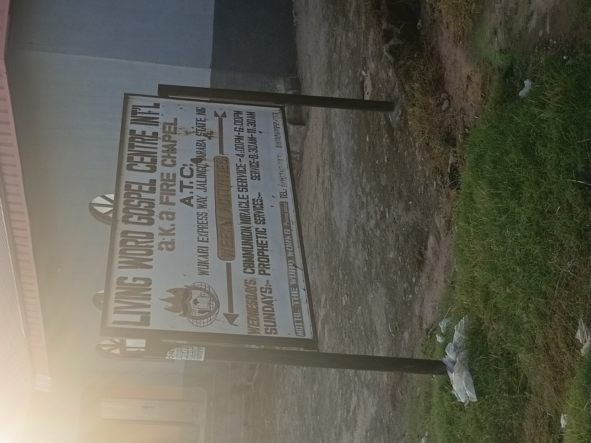 The image shows a sign for "LIVING WORD GOSPEL CENTRE INTL" located in Jalingo, Nigeria. The sign is white with black lettering and a logo. It indicates that the church is also known as "a.k.a FIRE CHAPEL A.T.C." and is situated on Wukari Express Way, Jalingo, Taraba State, Nigeria. The sign lists weekly activities including "COMMUNION/MIRACLE SERVICE" on Wednesdays from 4:00 PM to 6:00 PM, and "PROPHETIC SERVICES" on Sundays from 8:30 AM to 11:30 AM. A motto, "THE WORD WORKS," is also visible, along with contact information. The sign is supported by two metal poles. The surrounding environment appears to be an outdoor area with dry ground and some grass, possibly near a road or building. There is some plastic debris in the grass near one of the signposts. The lighting suggests it might be daytime, with a bright glare from the sun in the upper left portion of the image.