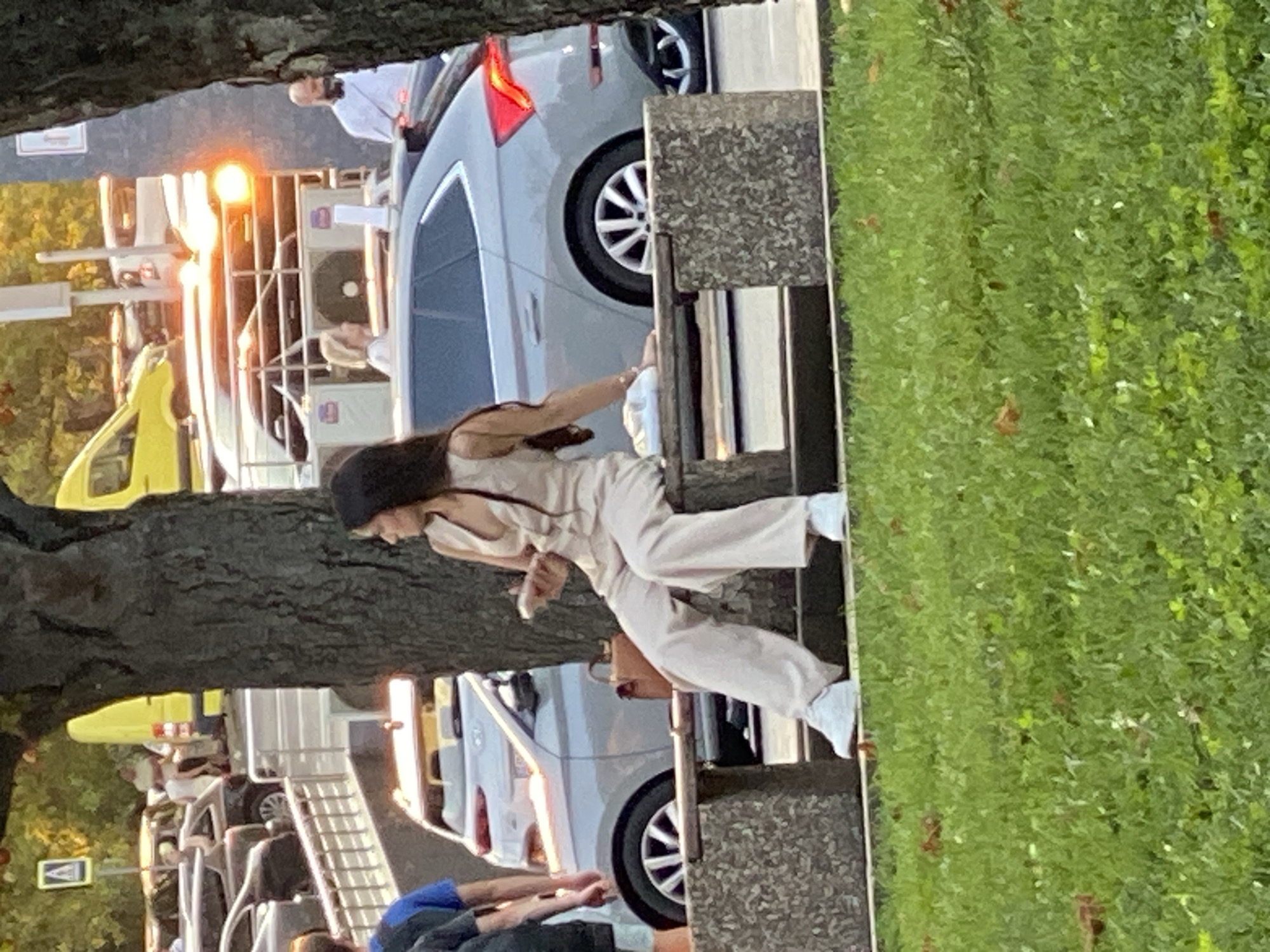 A woman with long, dark hair, wearing a light-colored sleeveless jumpsuit and white sneakers, sits on a stone bench. She is holding a mobile phone in her right hand and appears to be looking away from the camera. Next to her on the bench is a light-colored bag. In the background, a light gray hatchback car is parked. Behind the car, other vehicles and people are visible, along with trees and a yellow vehicle. The scene takes place outdoors, with a grassy area in the foreground. The overall impression is of a casual moment in a public urban setting.