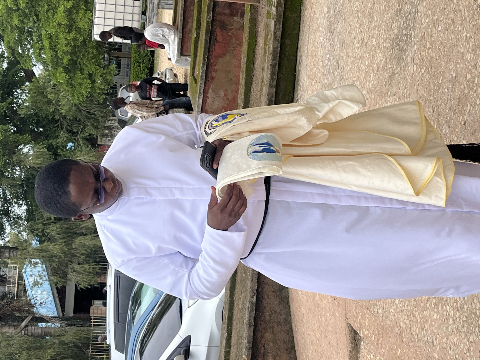 A Black man, likely a priest, is shown from the chest down, wearing a white robe with a black sash and glasses. He is holding a black smartphone in his right hand and a cream-colored garment with embroidered symbols in his left hand. The garment appears to be a religious vestment, possibly a chasuble, with a depiction of two figures on it.
The man is standing on a textured, reddish-brown paved surface, which could be a pathway or courtyard. In the background, several other people are visible. To the right, two young Black boys are seated on a raised, moss-covered stone structure that appears to be a tomb or monument. Another young Black man in a white robe is also seated on this structure. Further back, a group of young Black men are standing near parked vehicles, including a white car and a darker SUV.
Behind the people and vehicles, there is lush green foliage, including trees and bushes, suggesting an outdoor setting. A blue-roofed building with visible windows and architectural details is partially visible in the upper left quadrant of the image. The overall environment appears to be within a religious or educational institution, possibly the Vom Christian Hospital or a related facility, given the context of Vom, Nigeria, known for its medical and educational institutions. The lighting suggests it is daytime.