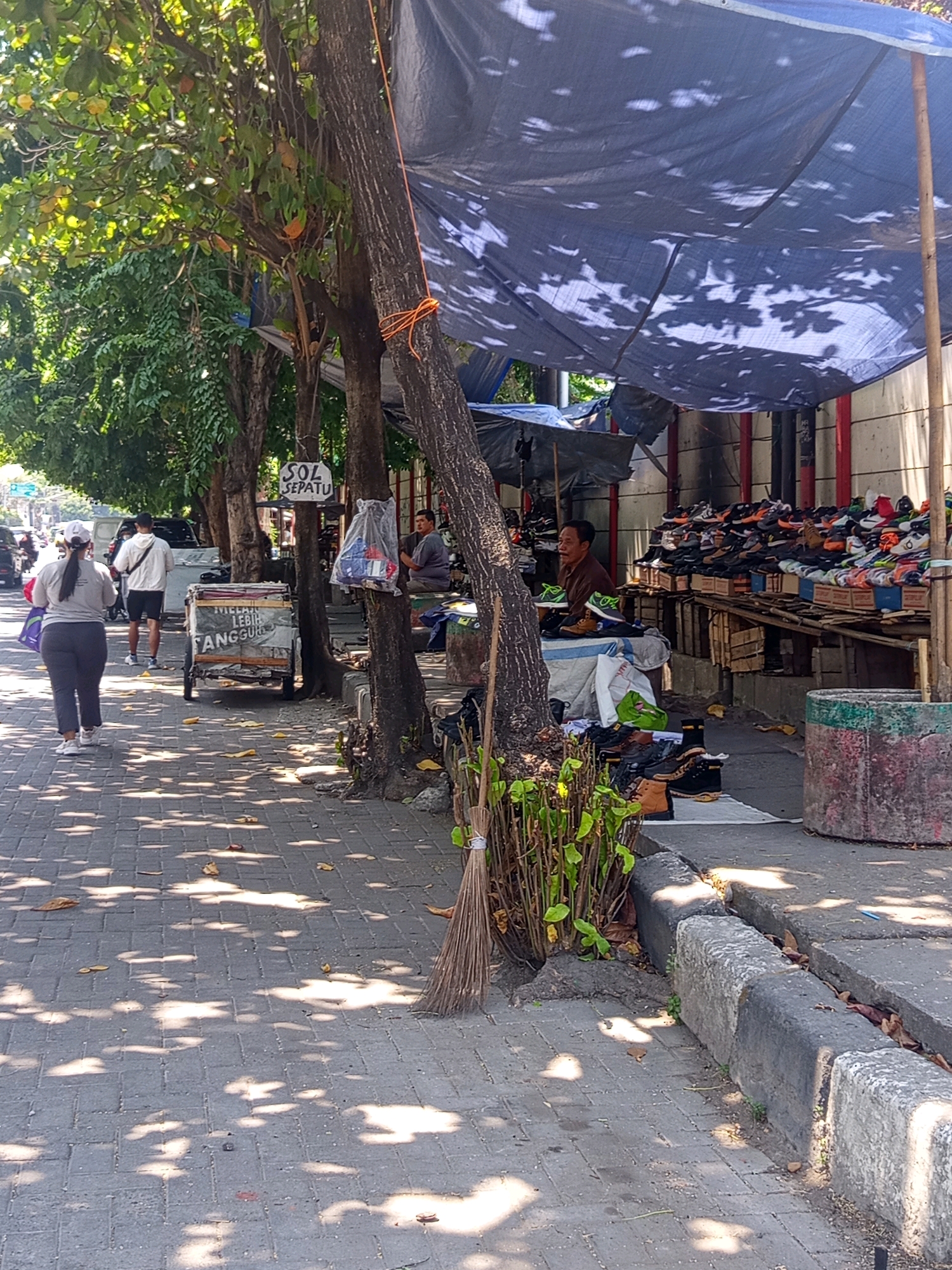 The image depicts a street scene in Semarang, Indonesia, focusing on a sidewalk shoe stall. A blue tarp, supported by wooden poles, provides shade over the stall. The stall displays a large variety of shoes arranged on wooden shelves and in boxes. A man, likely the vendor, sits behind the stall wearing a brown shirt. In the foreground, a woman wearing a white cap, grey t-shirt, and grey pants walks away from the camera. Another man, wearing a white jacket and black shorts, walks in the same direction ahead of her. To the left of the sidewalk, trees with green leaves cast dappled shadows on the brick-paved walkway. A small, white cart with writing on its side, reading "MELAN LEBIH TANGGUNG", is parked near the tree. To the right, a broom leans against a tree and a concrete planter sits on the edge of the sidewalk. The overall environment suggests a sunny day in a commercial area.