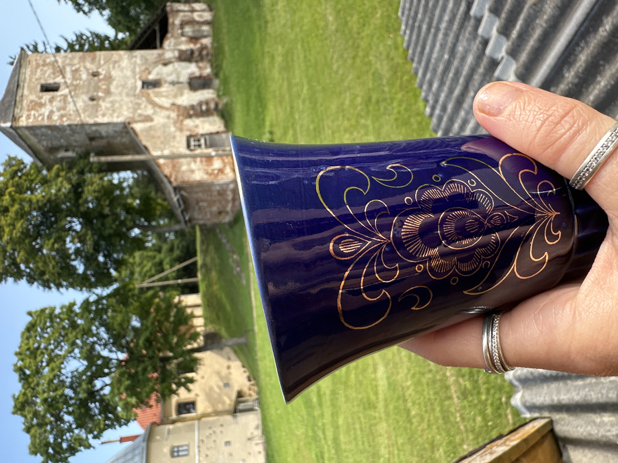 In the foreground of the image, a person's hand is holding a dark blue ceramic mug decorated with gold floral patterns. The mug is tilted, showing its opening and the design on its side. Two silver rings are worn on the person's fingers. In the background, a sunlit grassy area is visible, with several trees. To the right, a weathered, multi-story stone building with a square tower and arched doorways stands in disrepair. To the left, a lighter-colored building with a red-tiled roof and a small window is partially visible, and beyond that, another section of a stone structure. The scene appears to be located at the Norviliškės Manor in Norviliškės, Lithuania.