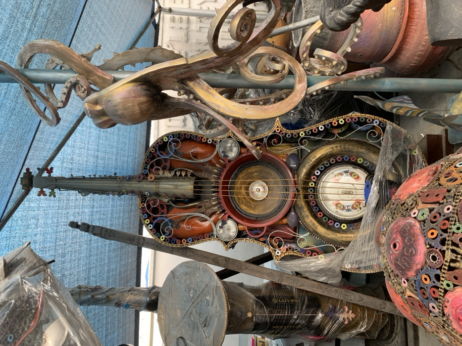 The scene depicts an outdoor art display or workshop area in Ikate, Nigeria. A large, approximately man-sized string instrument, resembling a cello or double bass, stands centrally, crafted from repurposed materials. Its body is primarily reddish-orange, intricately decorated with metallic gears, springs, bolts, and other industrial components, including two small headlights and a large speaker cone integrated into its soundboard area. The instrument's edges are adorned with numerous small, multi-colored rings. Below the speaker, a circular gold-colored frame surrounds a decorative white plate with patterns. To the immediate right, a large, multi-limbed sculpture, appearing metallic gold and bronze, resembles an octopus or abstract tendrils, affixed to a blue vertical support pole. Below this sculpture, several reddish-brown pottery items are visible. To the left, a tall, cylindrical object wrapped in clear plastic stands next to a dark, vertically oriented carved wooden staff. In the foreground, bottom left, a dome-shaped sculpture is partially visible, covered in a mosaic of small, colorful metallic rings and red painted sections, also partially wrapped in clear plastic. A light blue tarp provides overhead cover for the area. No individuals are present in the image.