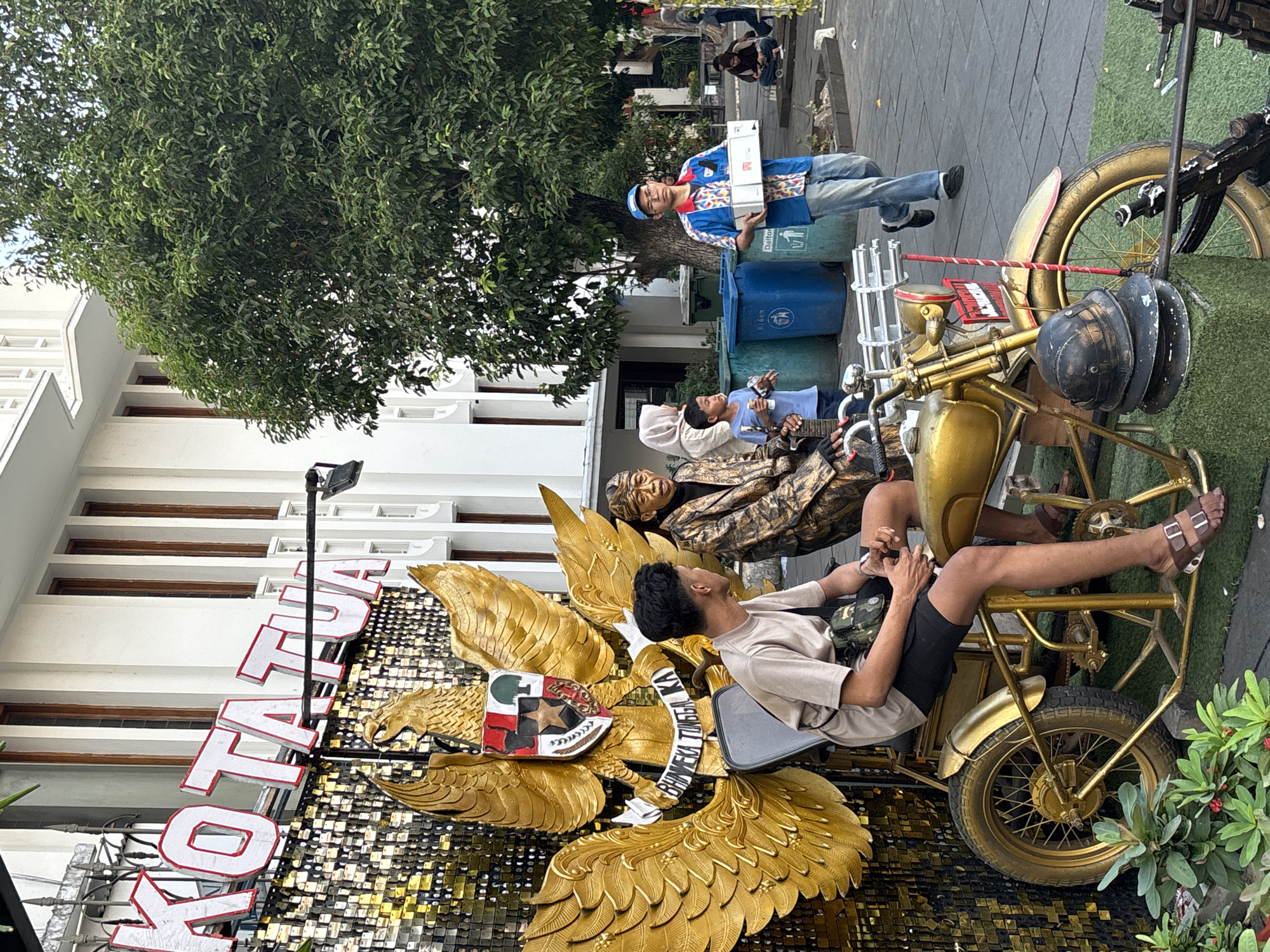 In the city of West Jakarta, Indonesia, a street scene unfolds with a prominent golden eagle emblem as the focal point. Two men, one young and casually dressed in a t-shirt and shorts, and an older man in a patterned jacket and headwear, are seated on a golden motorcycle. The younger man is on the left, facing to the right, while the older man is on the right, looking forward. Behind them, a large golden eagle statue with outstretched wings is mounted on a sequined backdrop that spells out "KOTA TUA" in large red letters. To the right of the motorcycle, a man in a blue shirt and cap walks carrying two white boxes. Several blue and green trash bins are visible in the background, along with a large green tree. The ground is paved with grey cobblestones, and some greenery is present in the foreground.