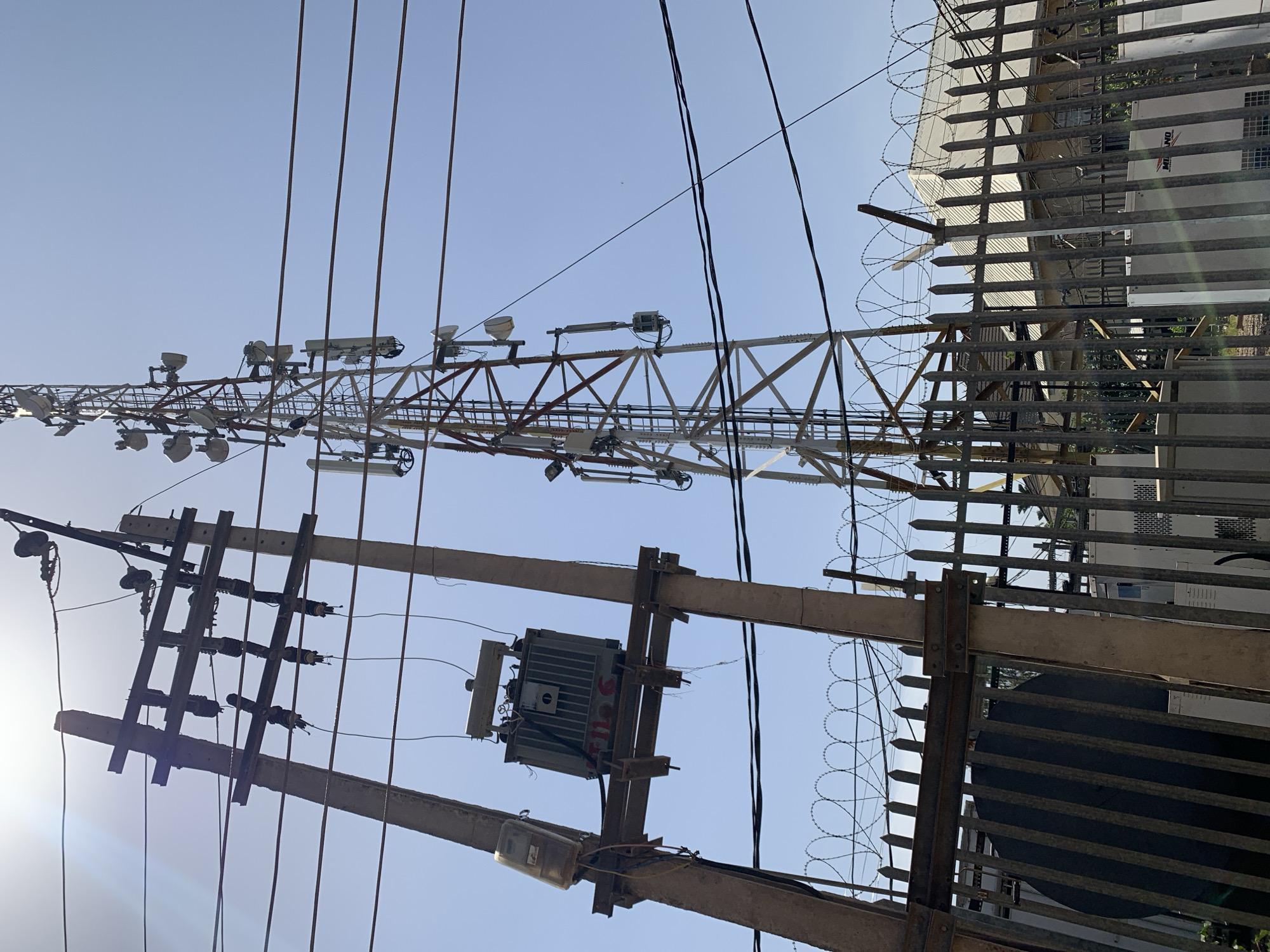 The image shows a low-angle view of utility infrastructure against a bright sky, possibly located in Rafin Par, Nigeria. In the foreground, there's a utility pole with a transformer and a meter attached. Electrical wires run from the pole. To the right, a metal fence topped with barbed wire runs vertically alongside the utility pole. In the background, a lattice tower is partially visible. The buildings partially blocked with the metal fence are white with windows.