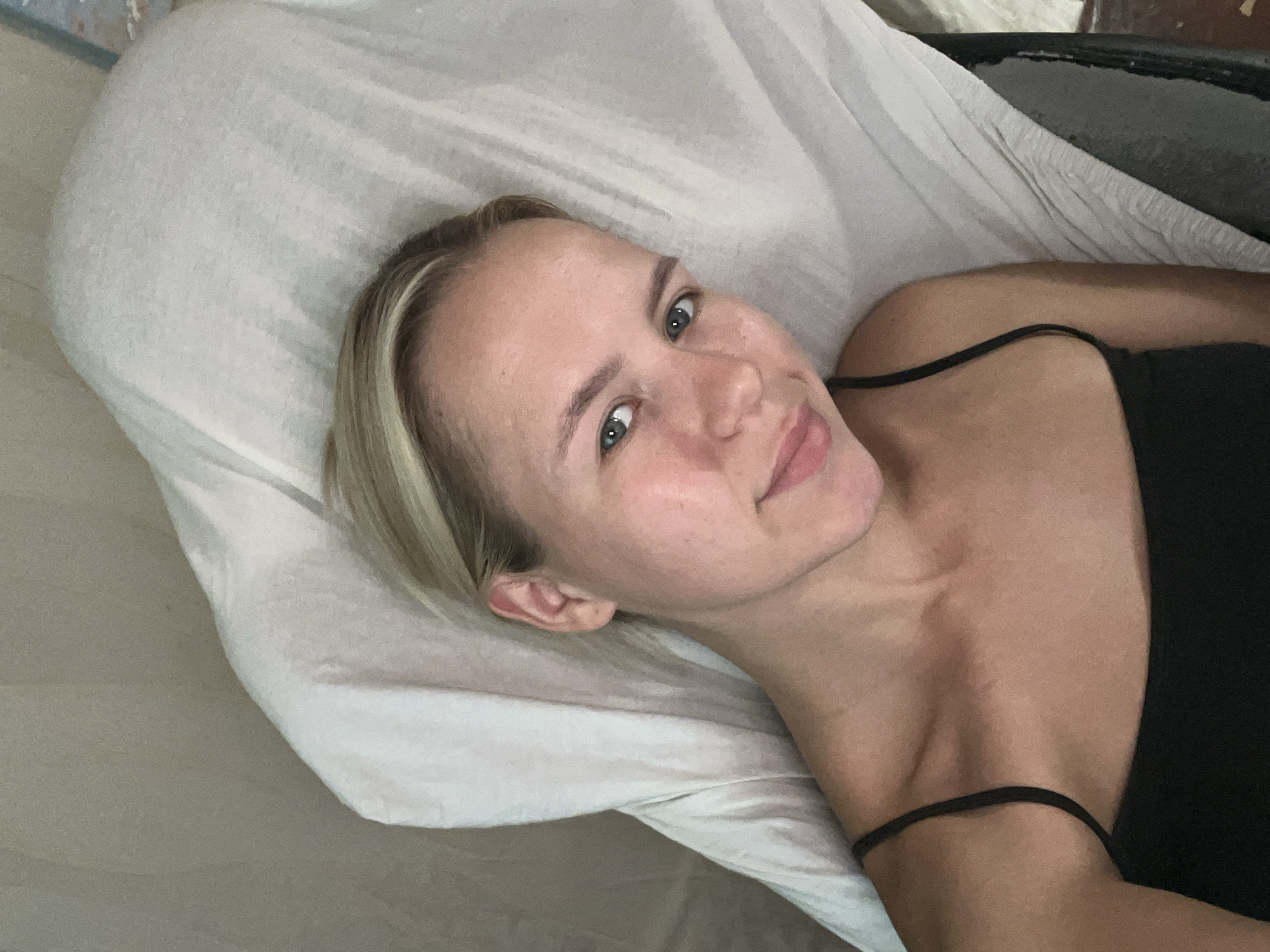 A young Caucasian woman with blonde hair is lying down on a white sheet with a grey wall in the background. She is wearing a black tank top. The woman is looking up at the camera and smiling. Her face is clean and clear, and her eyes are blue. The lighting is soft and natural.