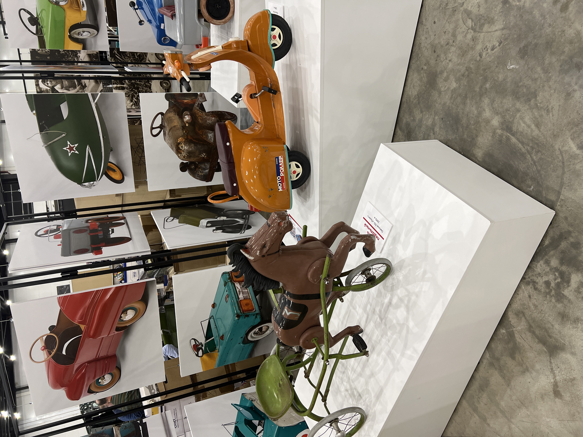 The image shows a display of vintage toy vehicles and photographic prints in an indoor exhibition setting, possibly in the city of Unknown, Unknown. The display includes a child's orange scooter with "MOTO РОДМЕР" printed on the side and a child's ride-on horse toy with a green metal frame and wheels. A black metal rack holds large photographic prints of similar vintage toy vehicles. The exhibition is set on a polished concrete floor. The scene is brightly lit, suggesting an indoor venue.