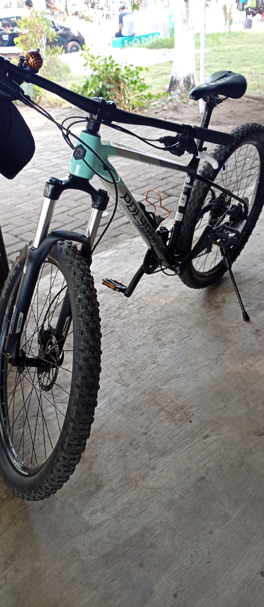 An outdoor, close-up, low-angle shot shows a light blue and gray mountain bicycle parked on a light gray concrete surface. The bicycle is positioned at an angle, with its front wheel on the left side of the frame and its rear wheel on the right. The front wheel is a knobby tire on a black rim. The bicycle has a suspension fork with silver stanchions and black lowers. The handlebars are black, with a small golden bicycle bell attached to the left side. A black bag or pouch is hanging from the left handlebar. The bicycle frame has "POLYGON" written in silver lettering. The rear wheel is also a knobby tire on a black rim. A black kickstand is extended, supporting the bike. A small orange bottle cage is attached to the frame. In the background, out of focus, a few people are visible, some seated on a blue bench. There is also greenery and trees, suggesting an outdoor area. The overall scene appears to be in a public area in Probolinggo, Indonesia, possibly a park or recreational space given the presence of people, trees, and a paved surface.